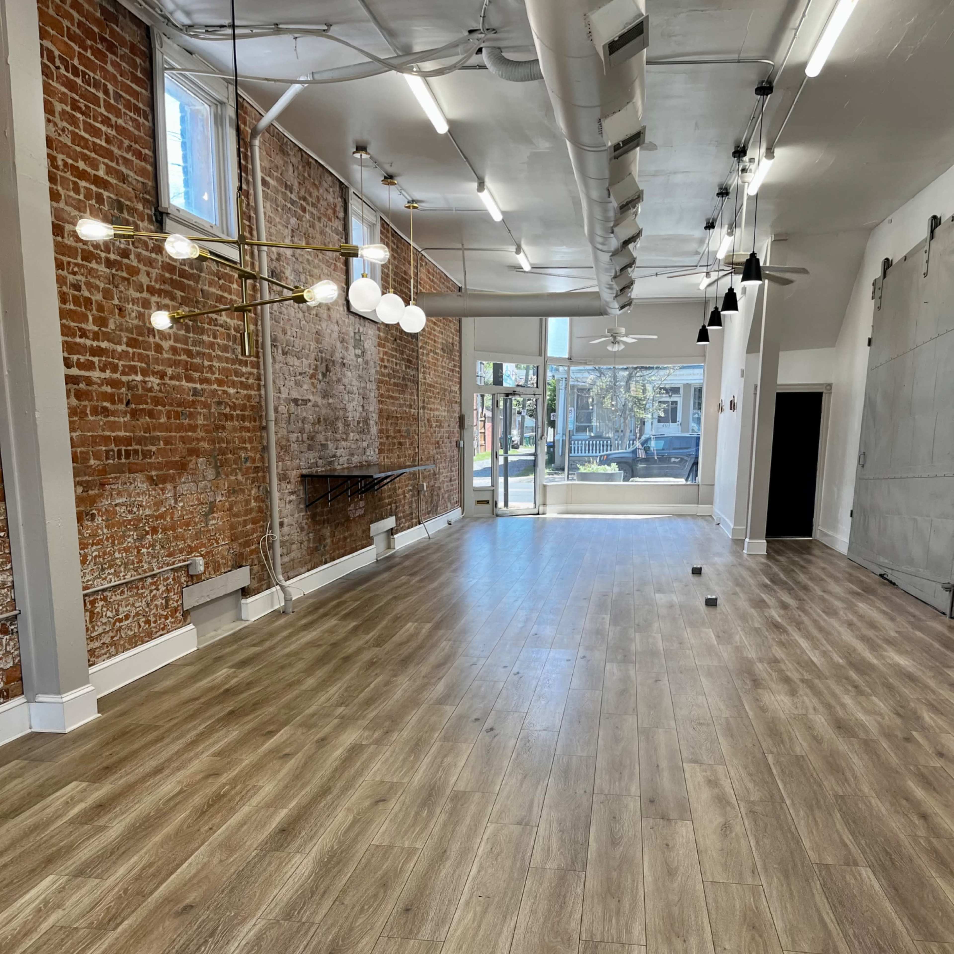 A spacious room with wooden flooring, exposed brick walls, and large windows allowing natural light, featuring modern light fixtures and ceiling fans.
