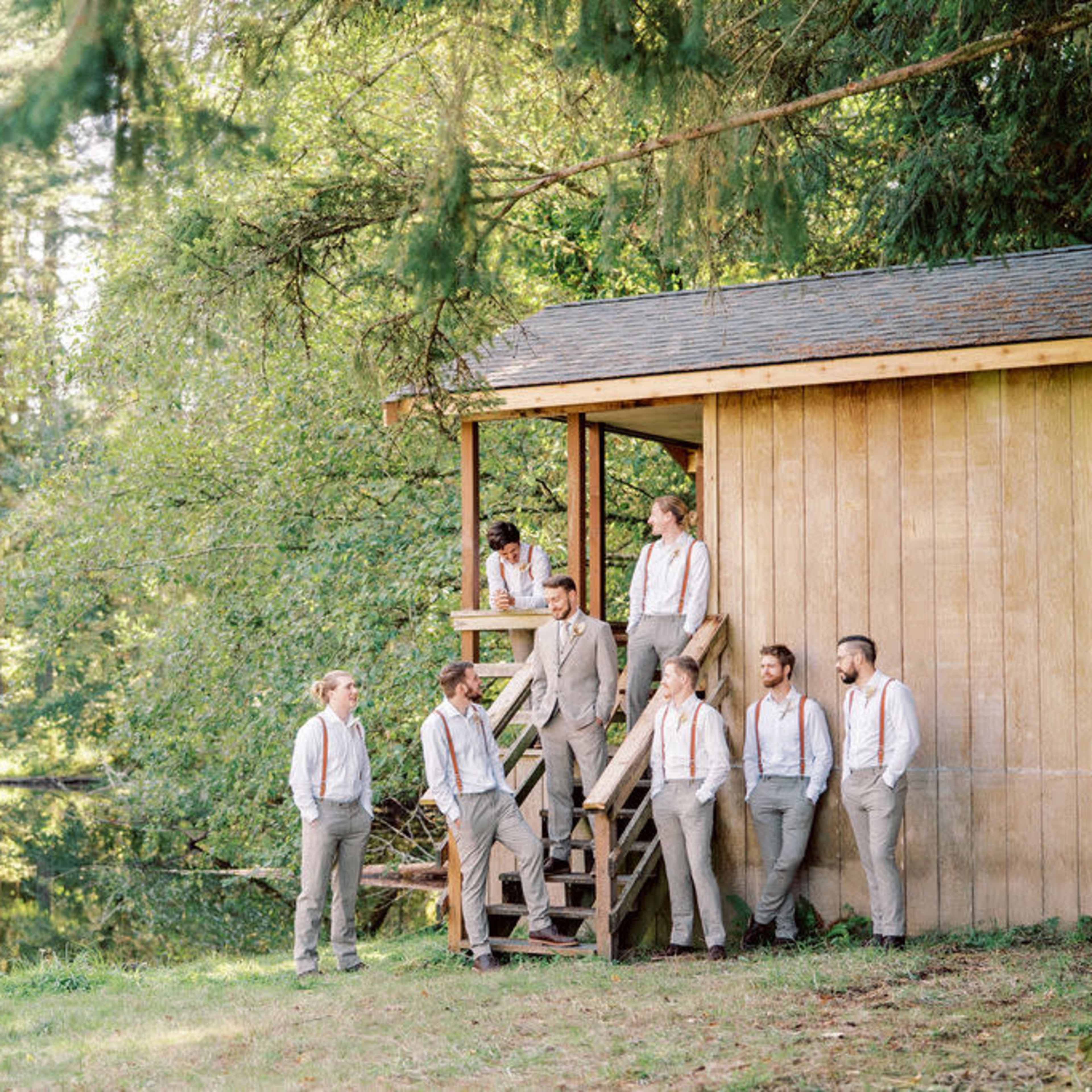 A group of eight men in light-colored suits and suspenders poses near a wooden cabin by a pond surrounded by trees.