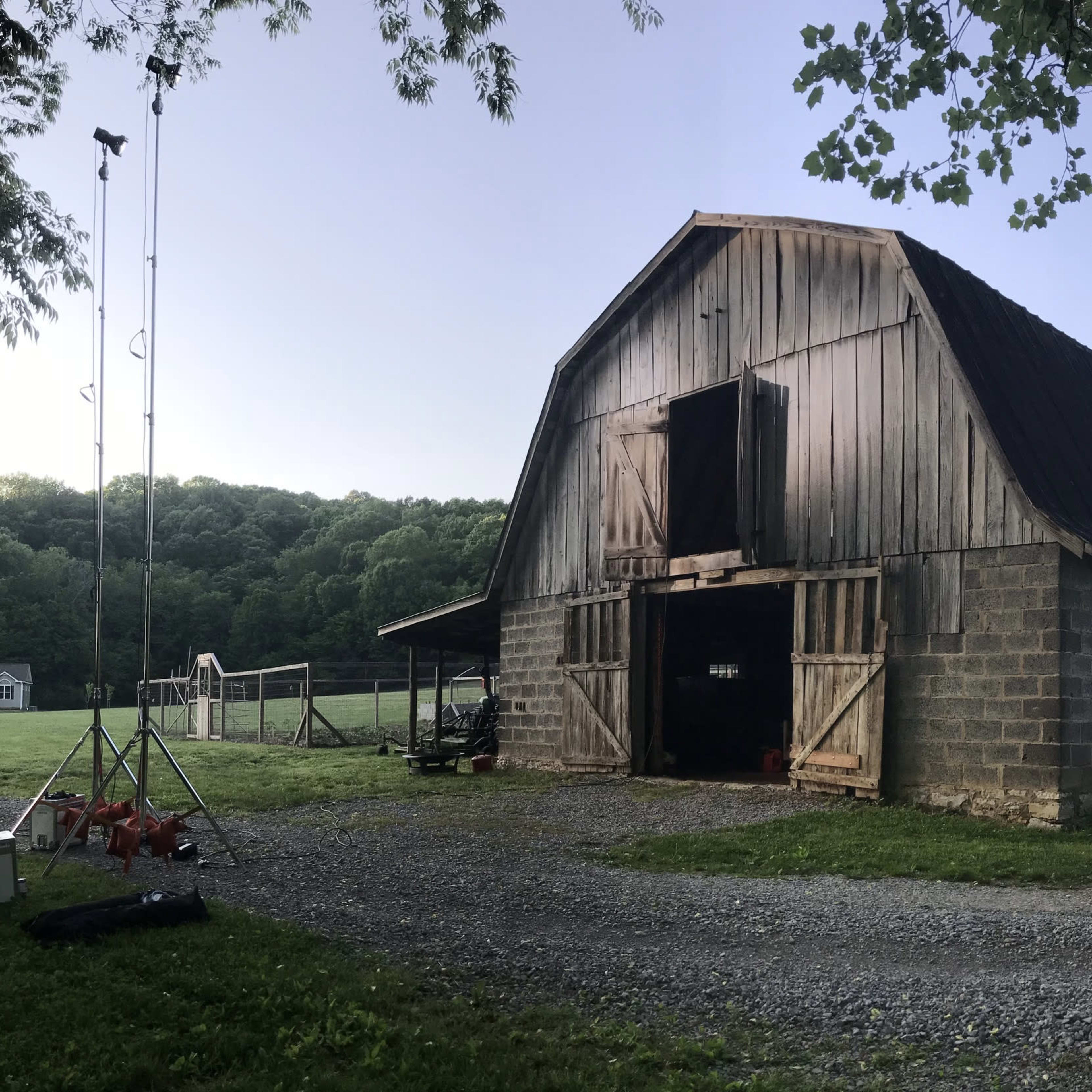 A large wooden barn with a metal roof stands beside a gravel path, surrounded by greenery and a field in the background.
