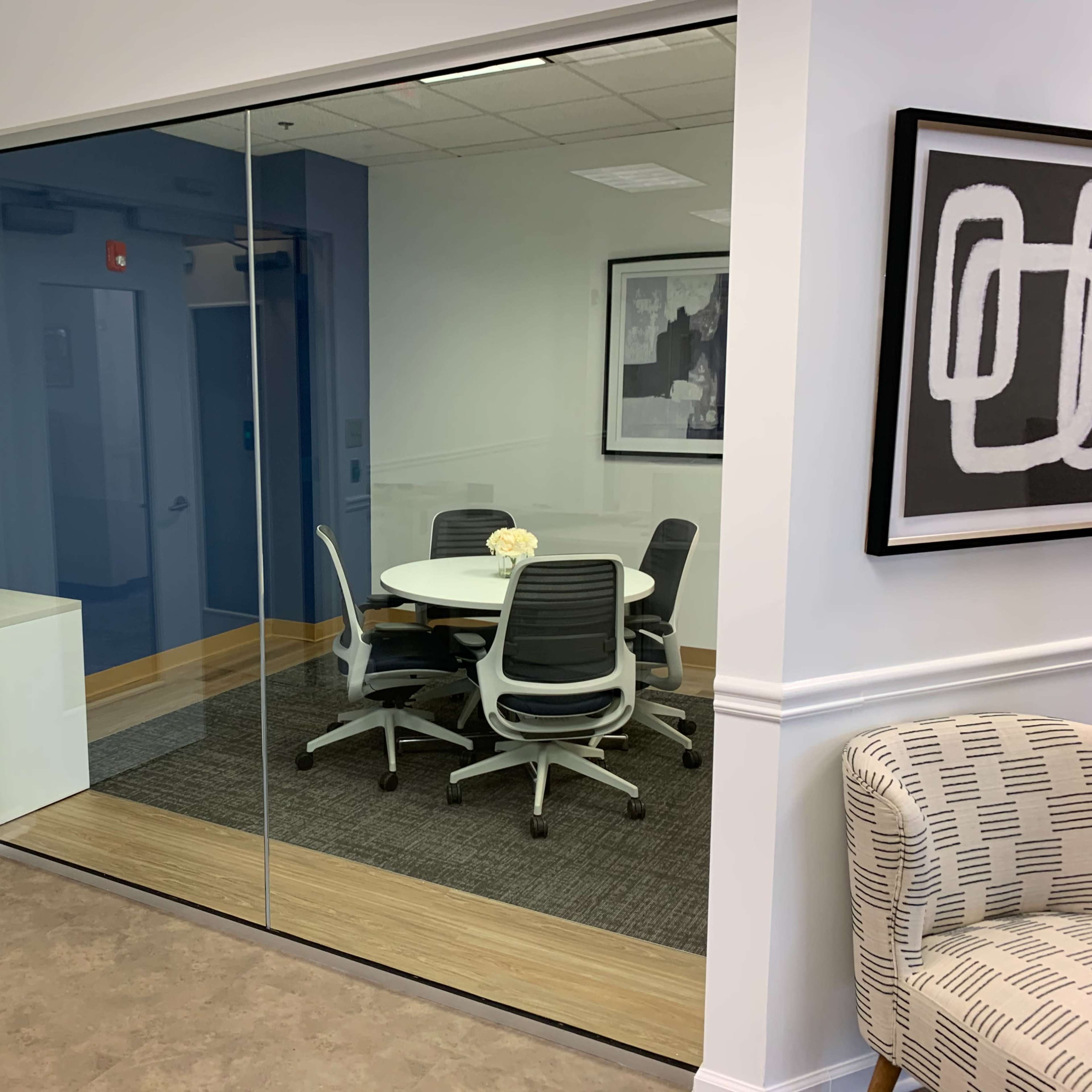 A modern conference room with a round table and five ergonomic chairs is visible through a large glass wall.