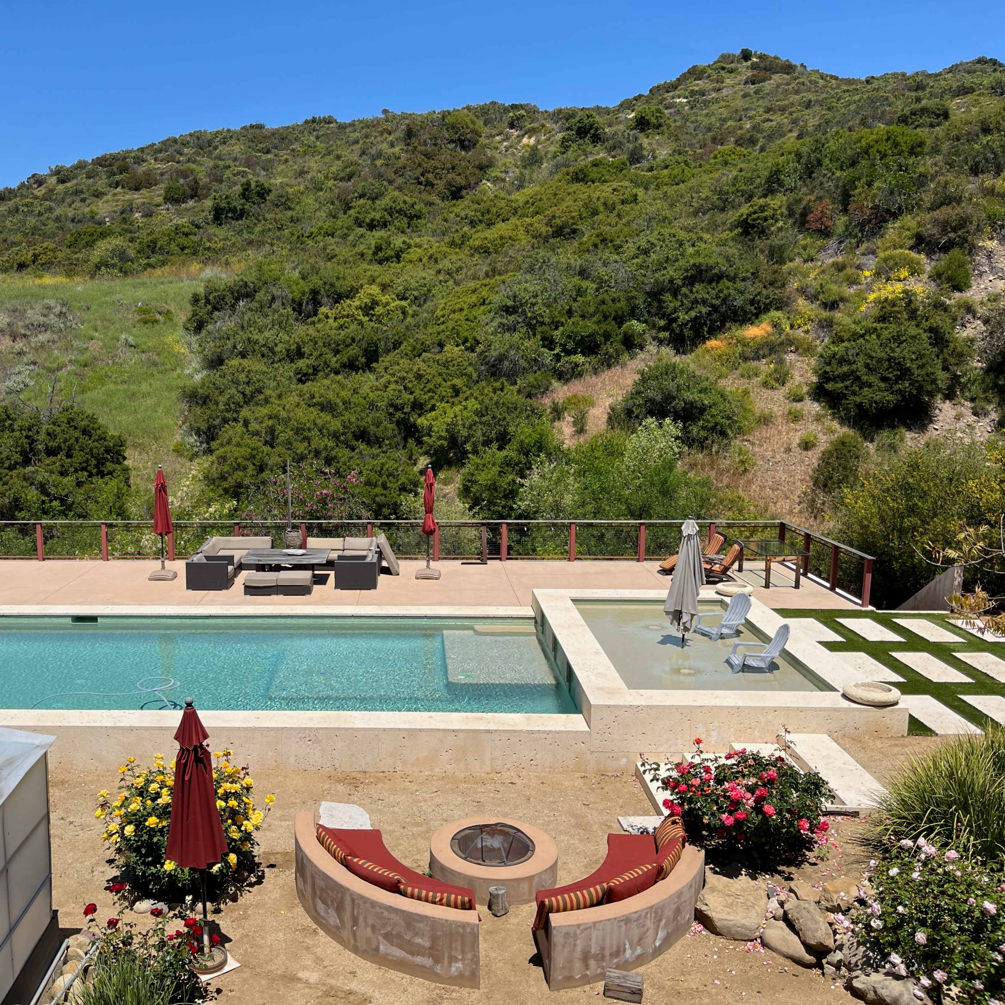 A backyard with a swimming pool surrounded by a deck, lounge chairs, and a seating area, set against a backdrop of lush green hills.