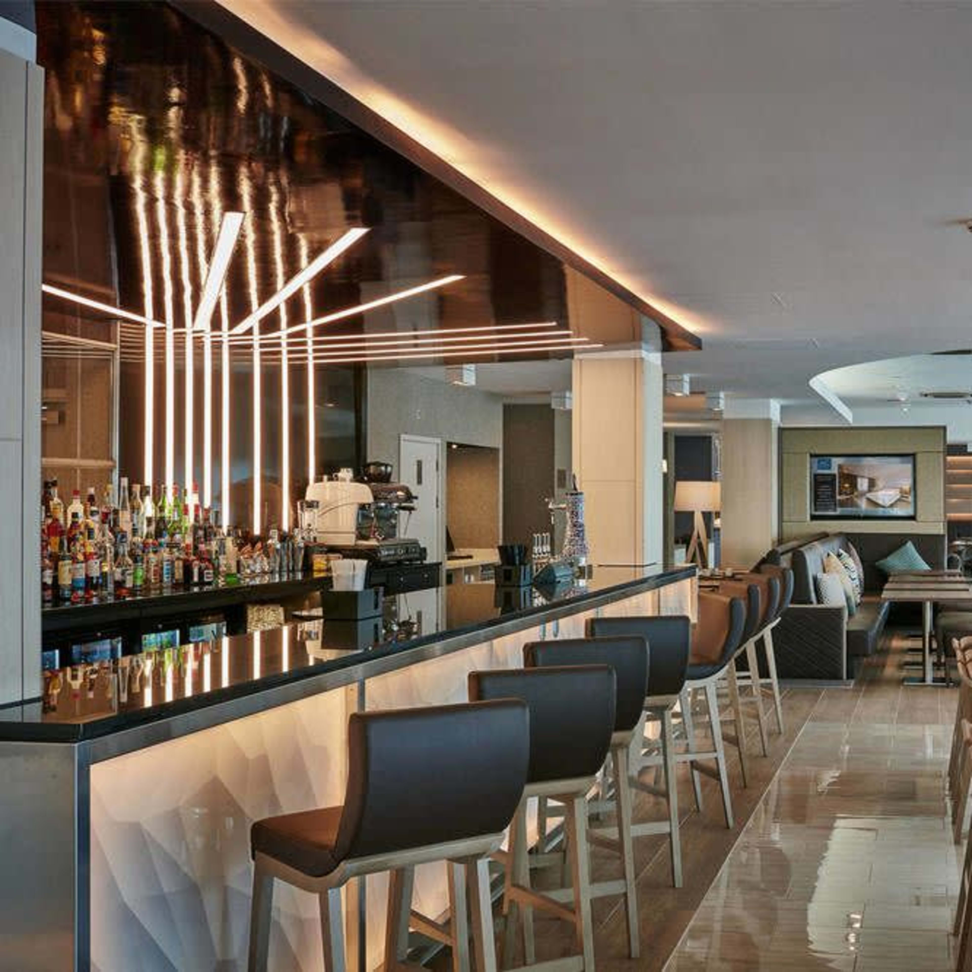 A modern bar area with a sleek counter, high stools, and a variety of bottles displayed on shelves.