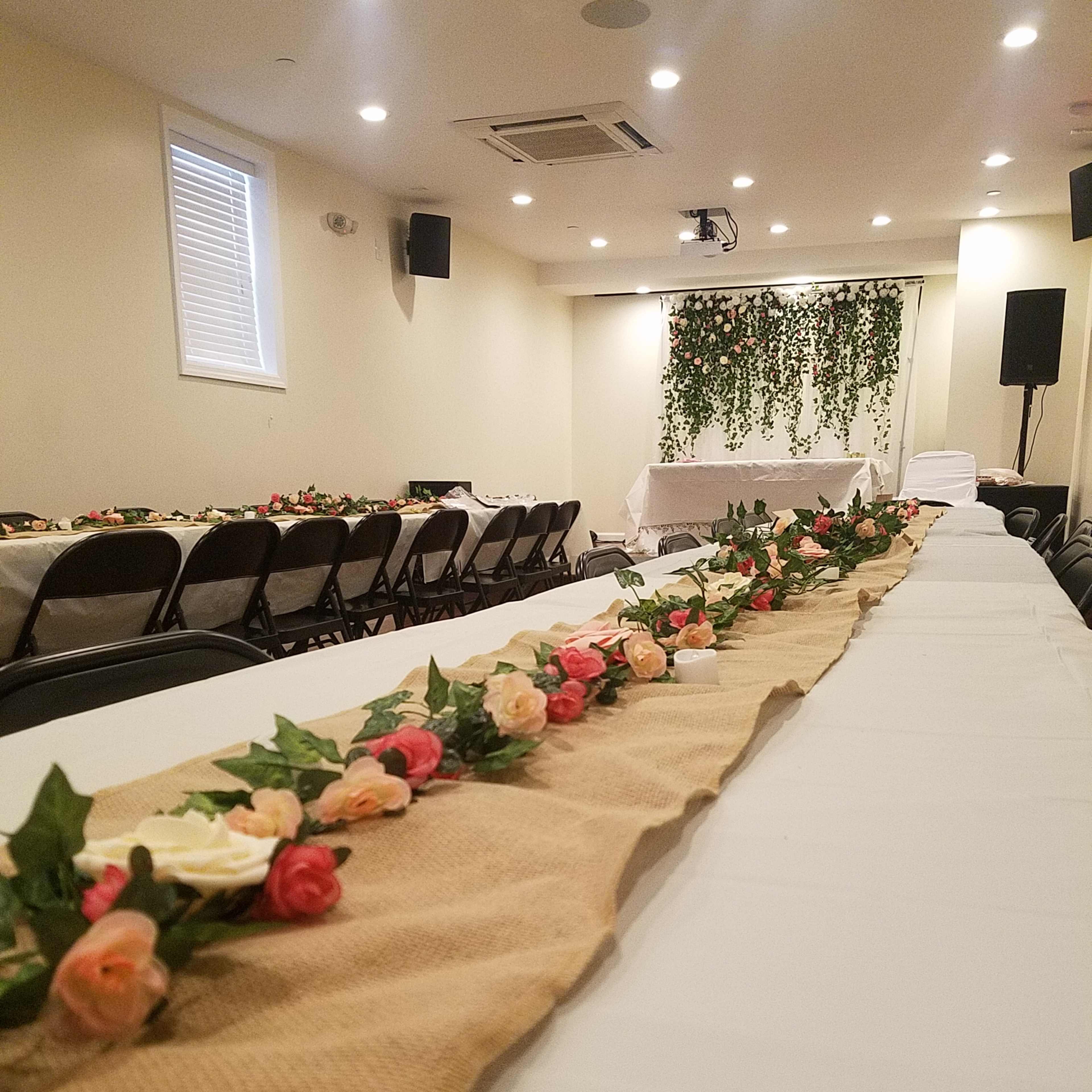 A banquet hall with long tables covered in white cloths, featuring a burlap runner adorned with artificial flowers, and a backdrop of greenery hanging on the wall.