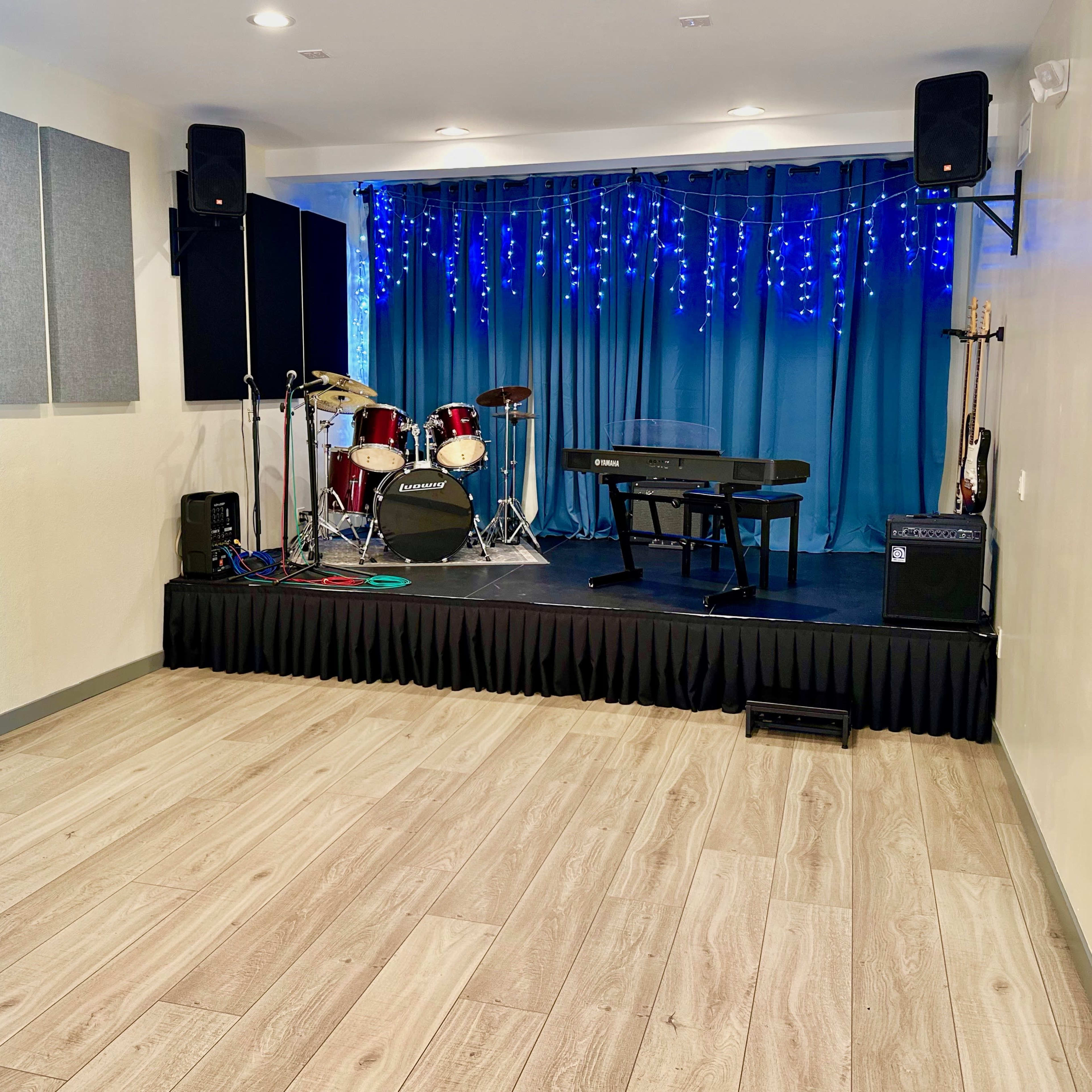 A small performance area featuring a stage with musical instruments, including a drum set and a keyboard, surrounded by blue curtains and string lights.