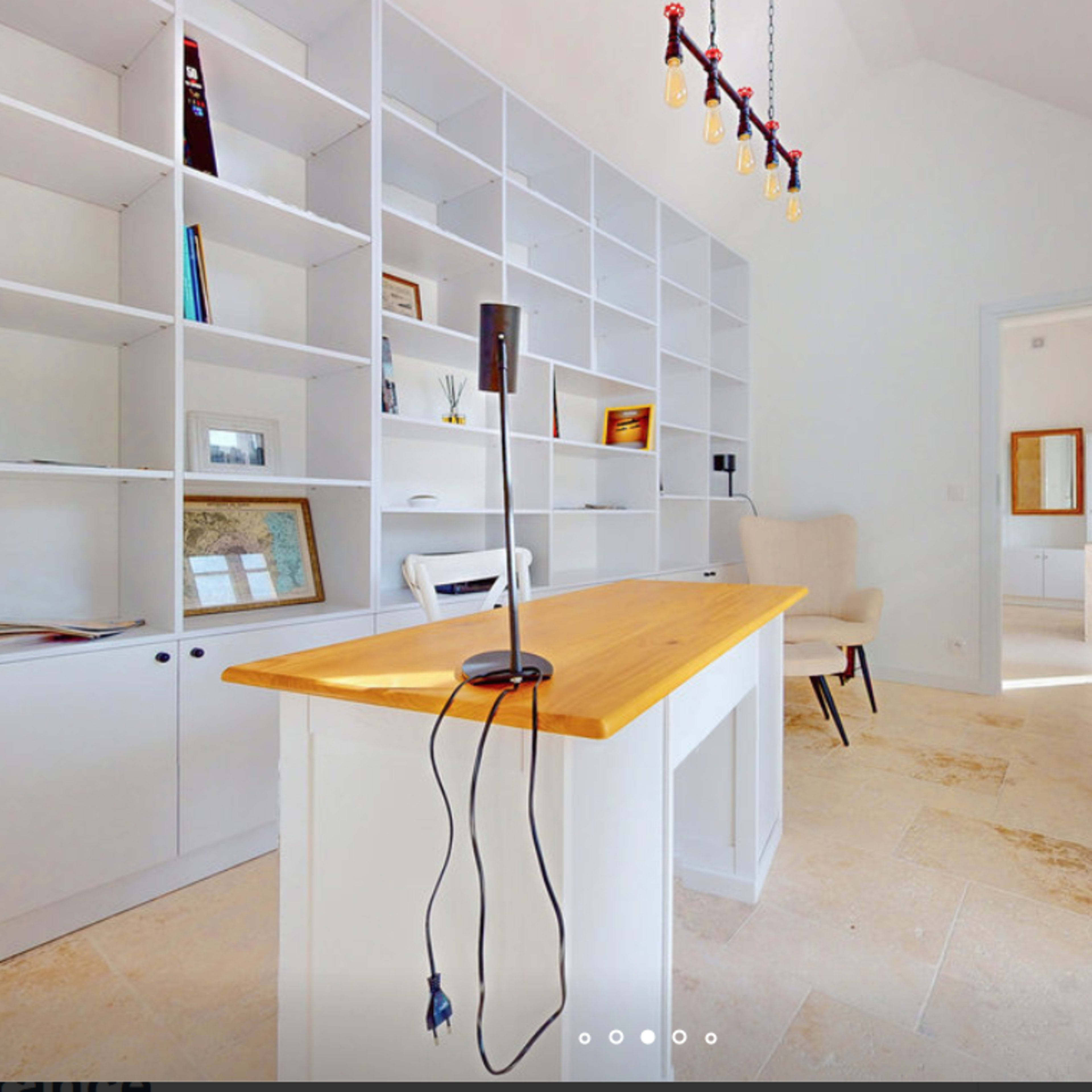 A modern room features a wooden table with a lamp, surrounded by white bookshelves and a doorway leading to another space.
