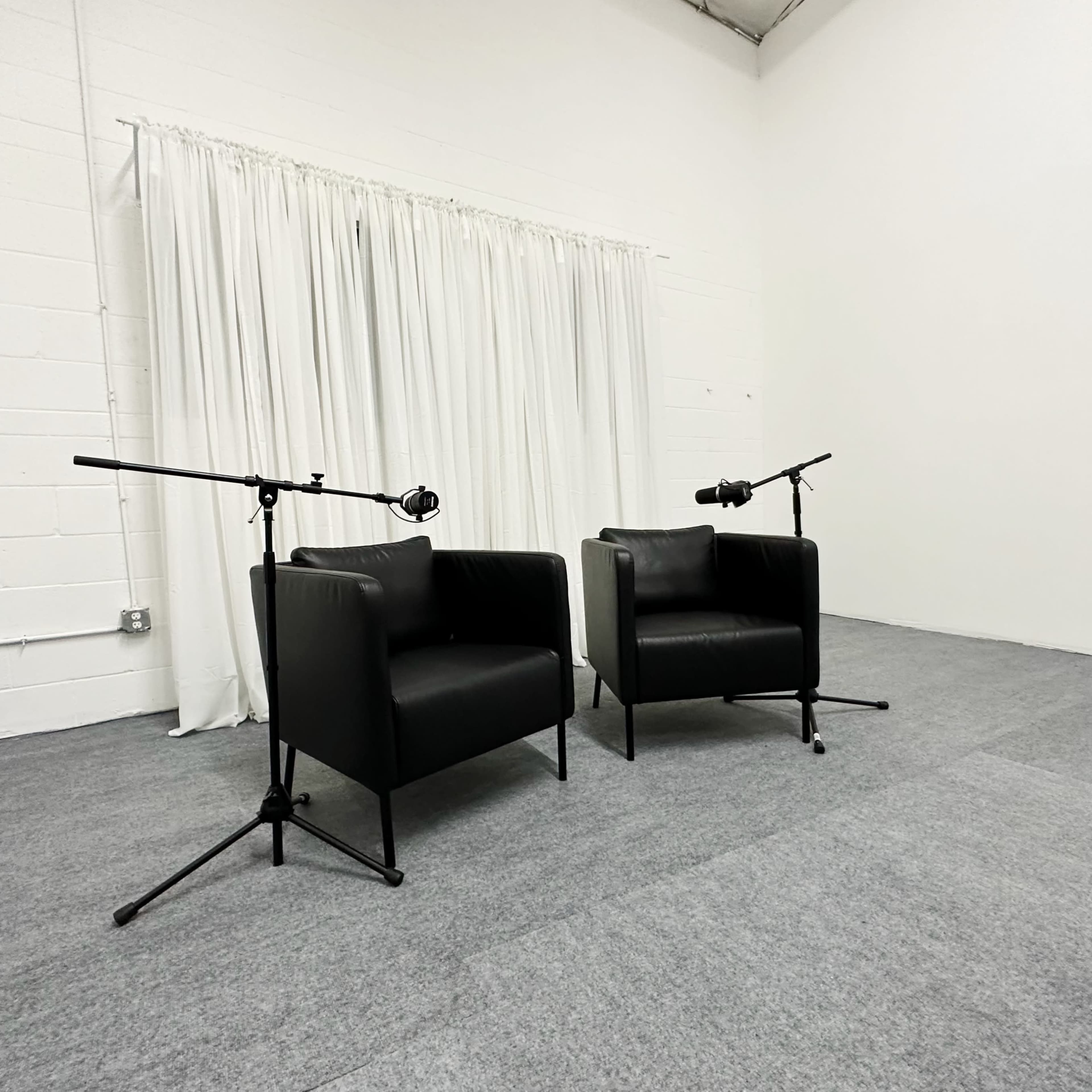 Two black chairs positioned across from each other, each outfitted with a microphone on a stand, set against a white curtain and gray carpet in a sparse room.