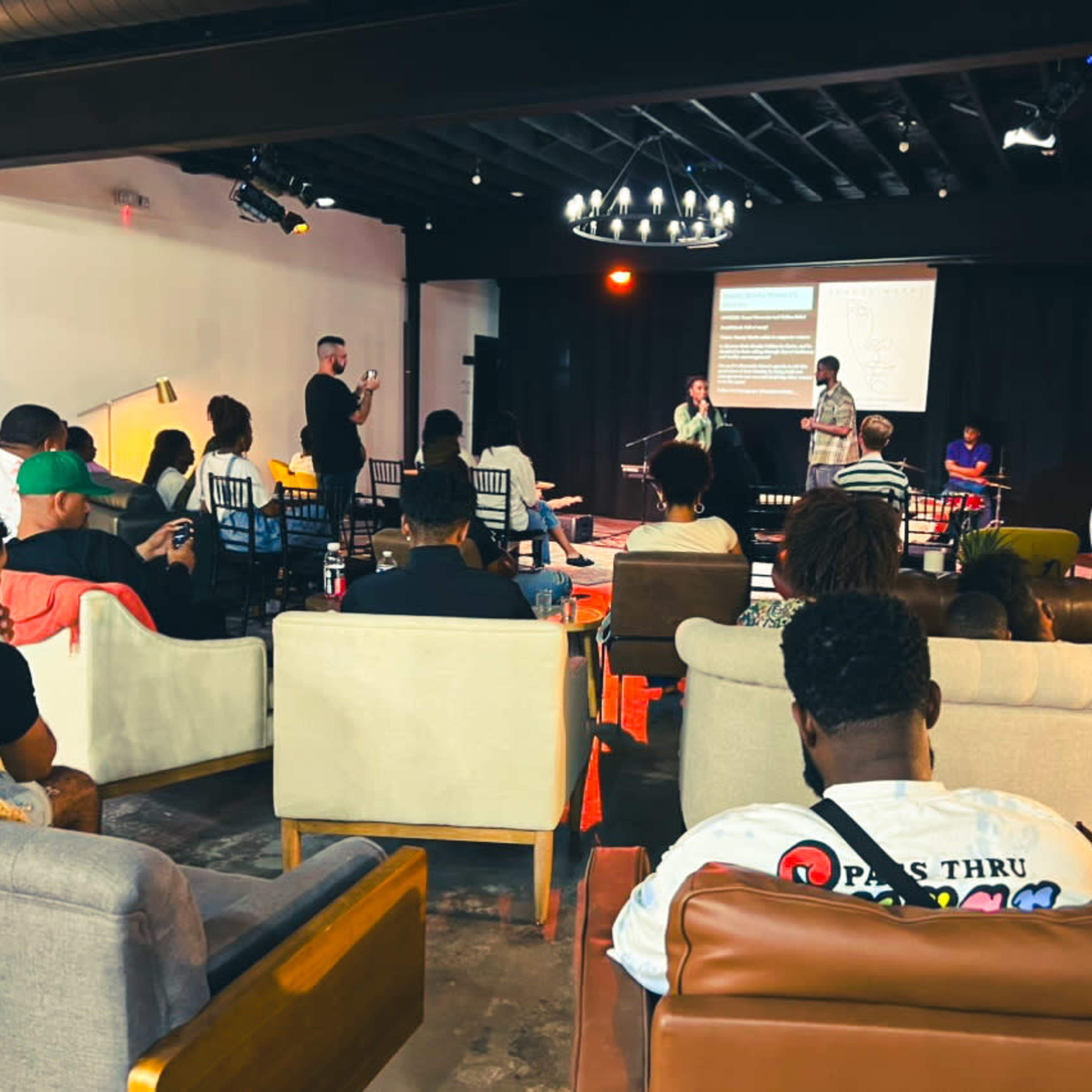 A group of people is seated in a lounge area while others stand and engage in a presentation on a stage in front of them.