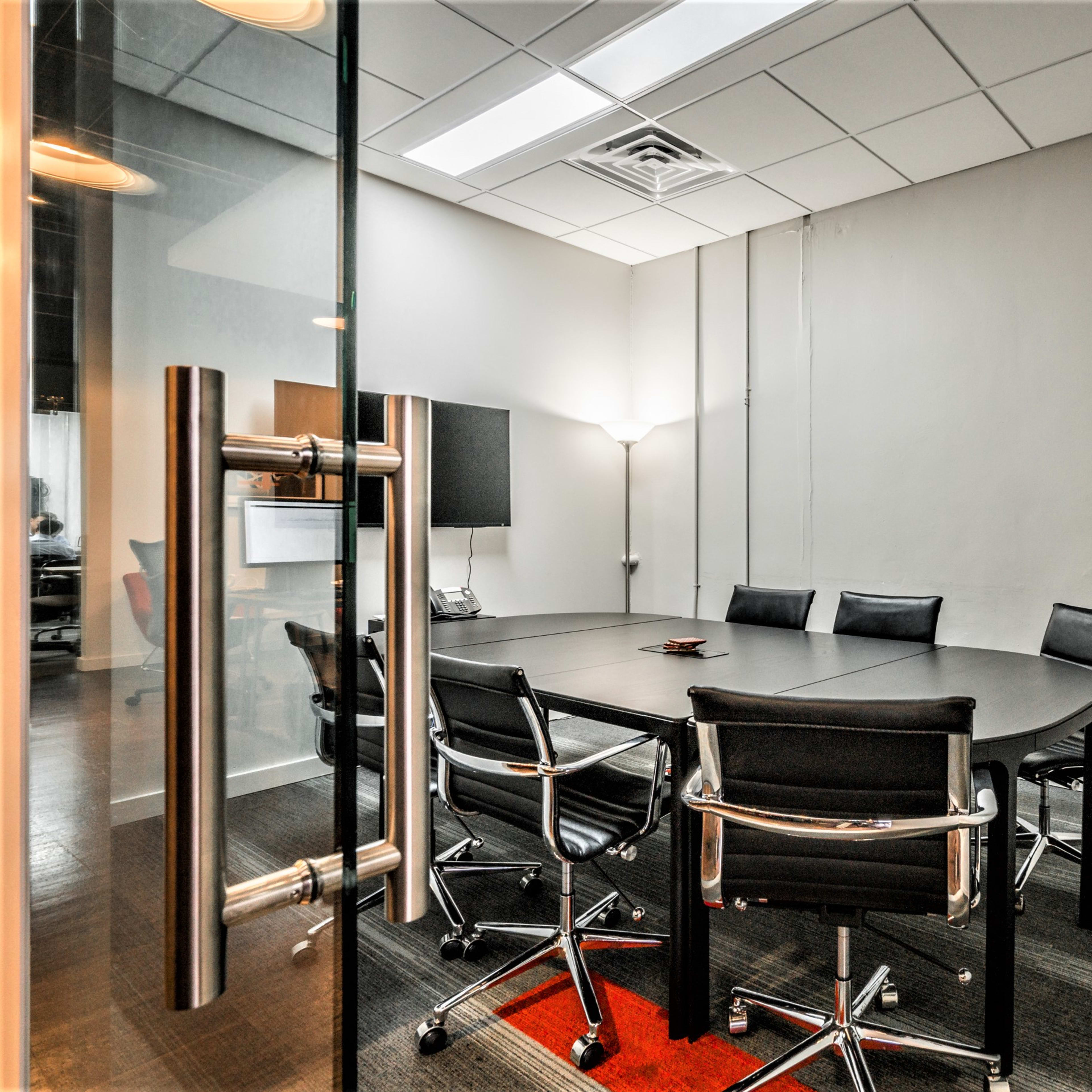 A conference room features a large round table surrounded by black chairs, with a television mounted on the wall and a lamp in the corner.