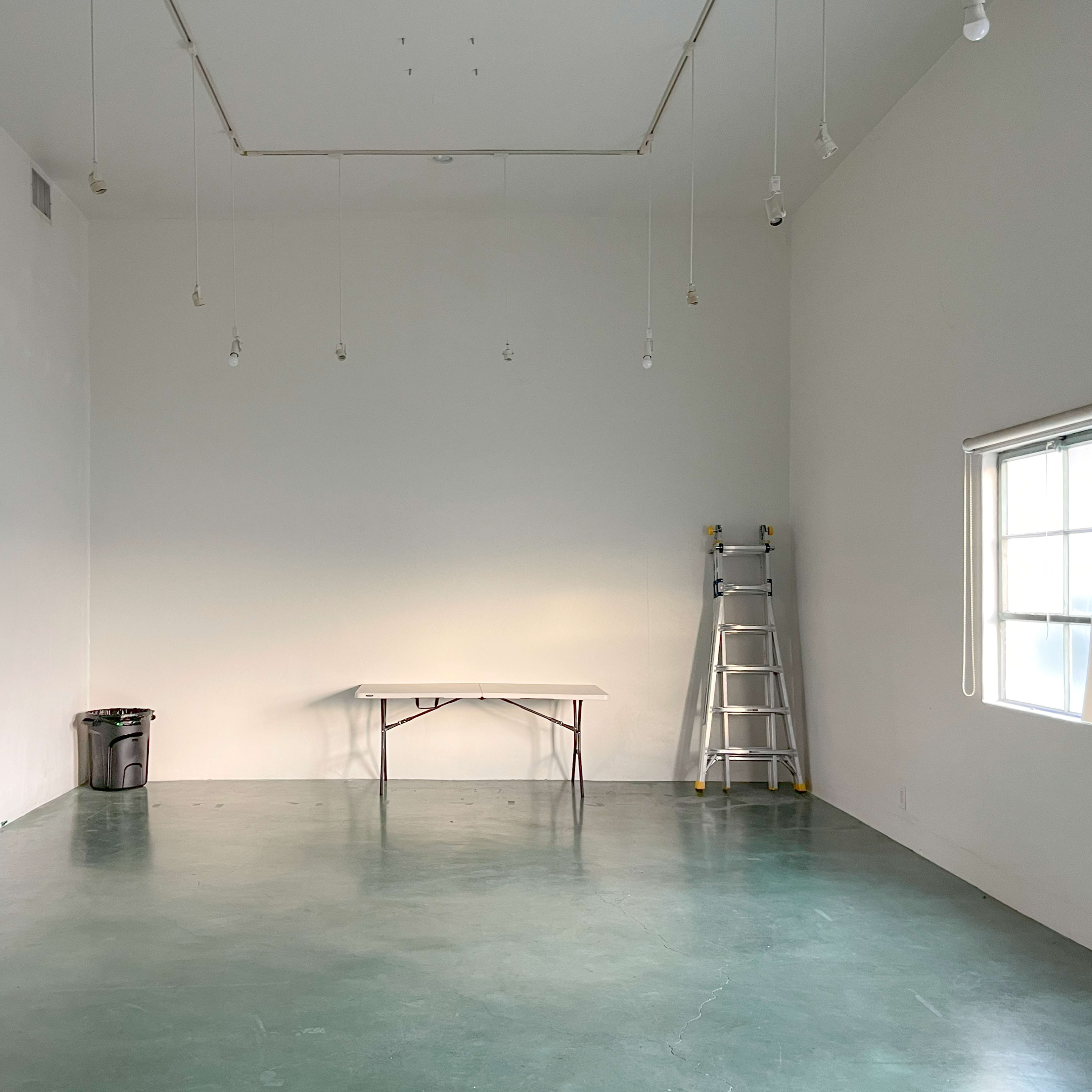 A large, empty room with a green concrete floor, a folding table in the center, a ladder against the wall, and a window on the right.
