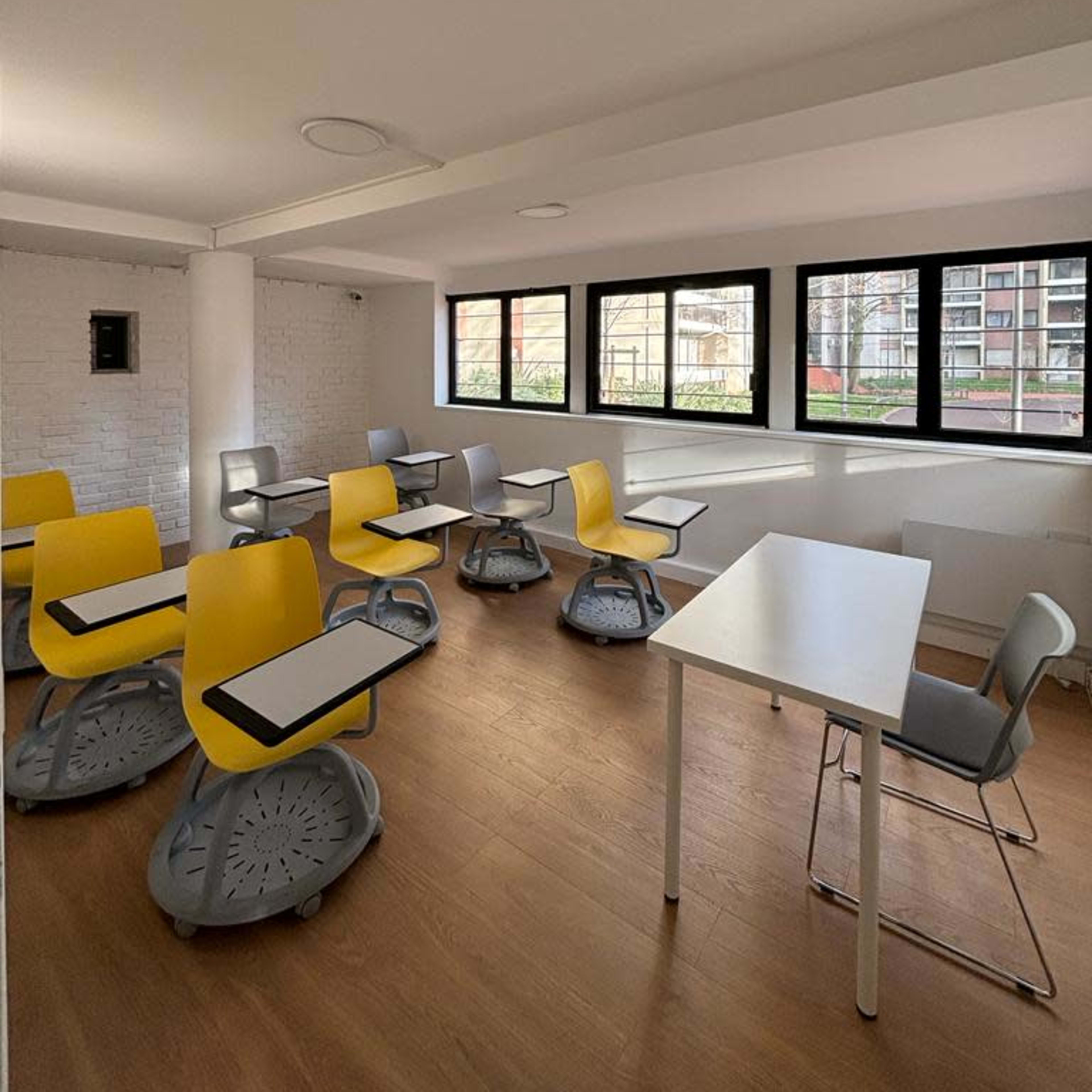 A bright classroom with several yellow and gray chairs arranged around white tables and large windows.