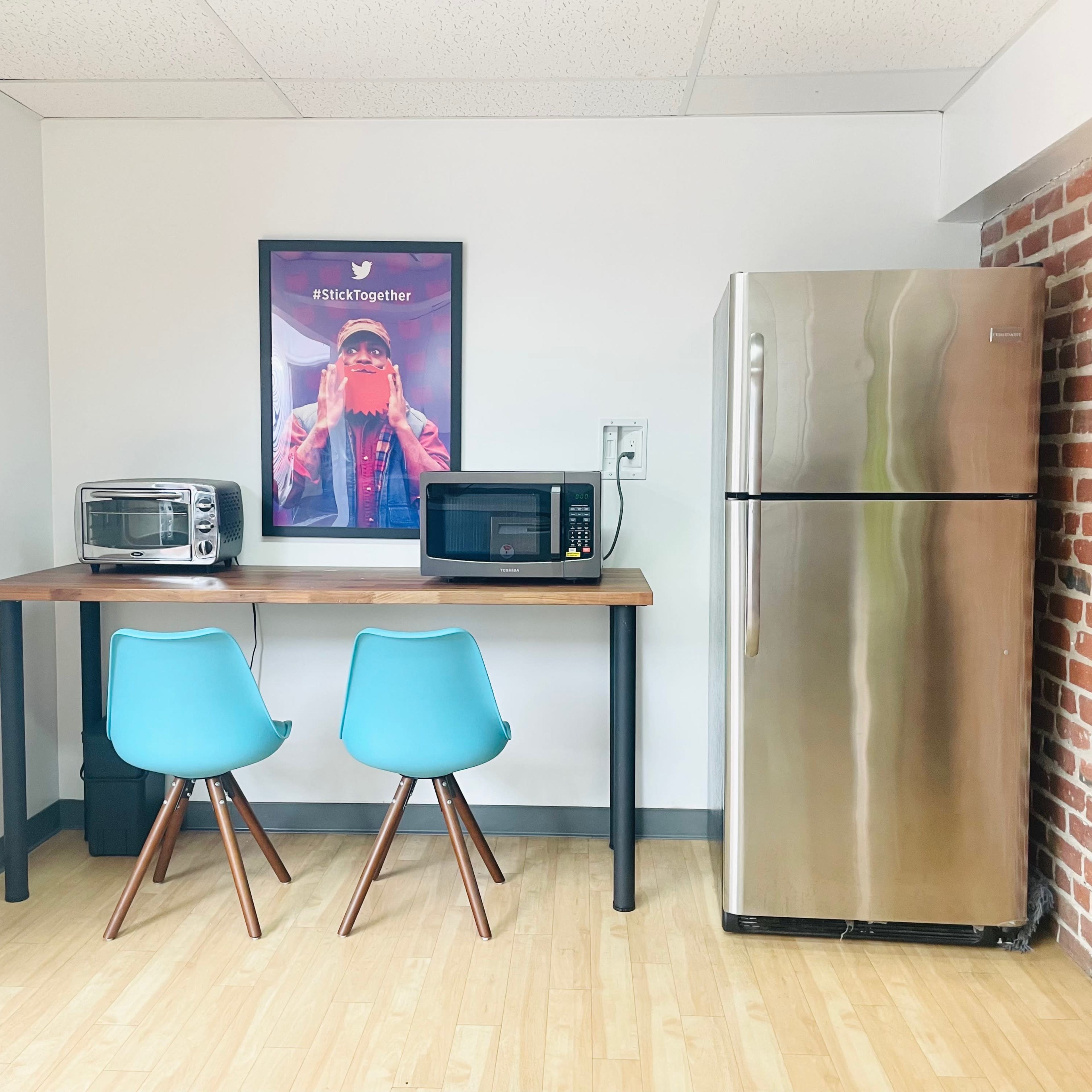 A small kitchen area featuring a wooden table with two blue chairs, a microwave, a toaster oven, and a large silver refrigerator against a brick wall.