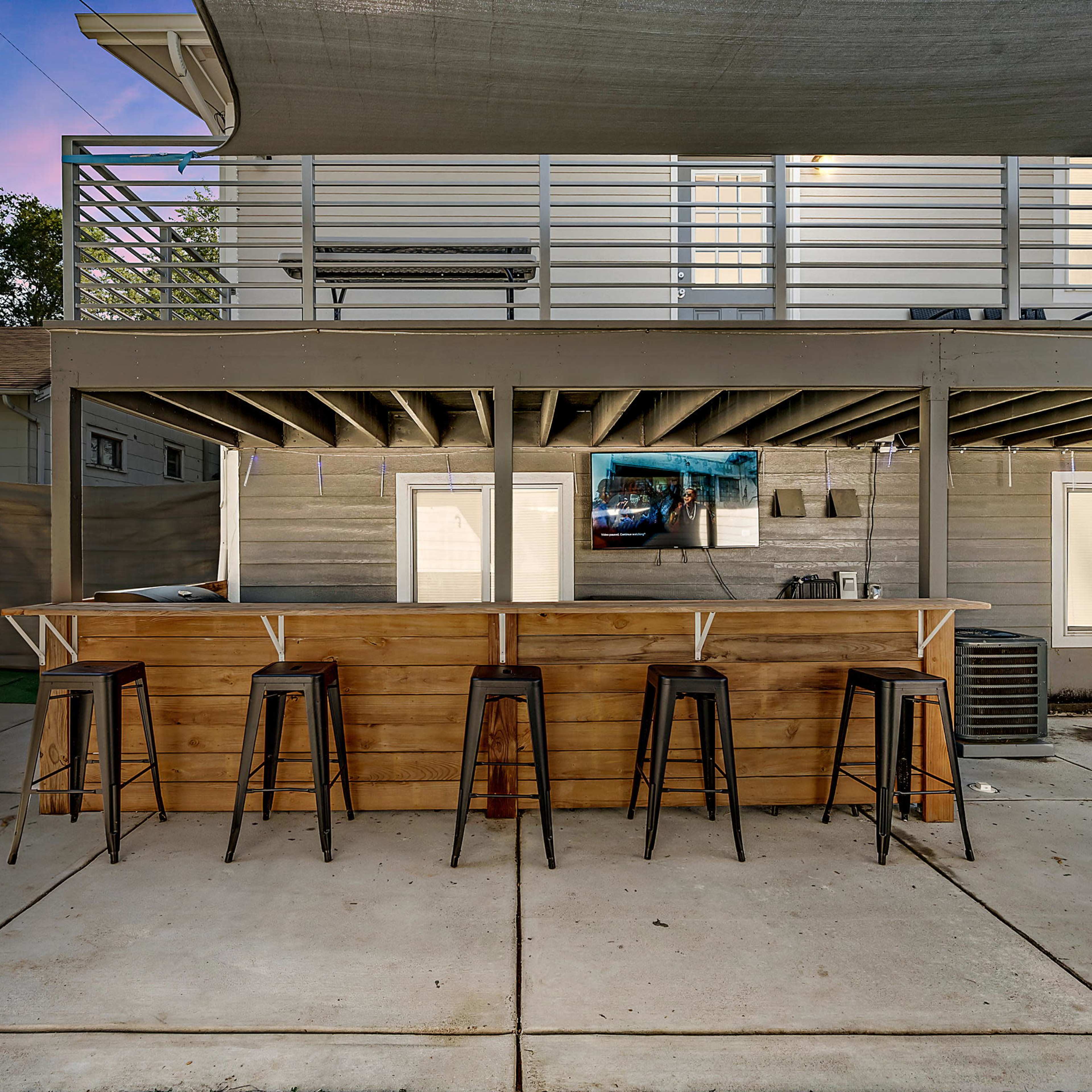 A modern outdoor bar area with a wooden counter and metal stools, set against the backdrop of a house with a covered patio.