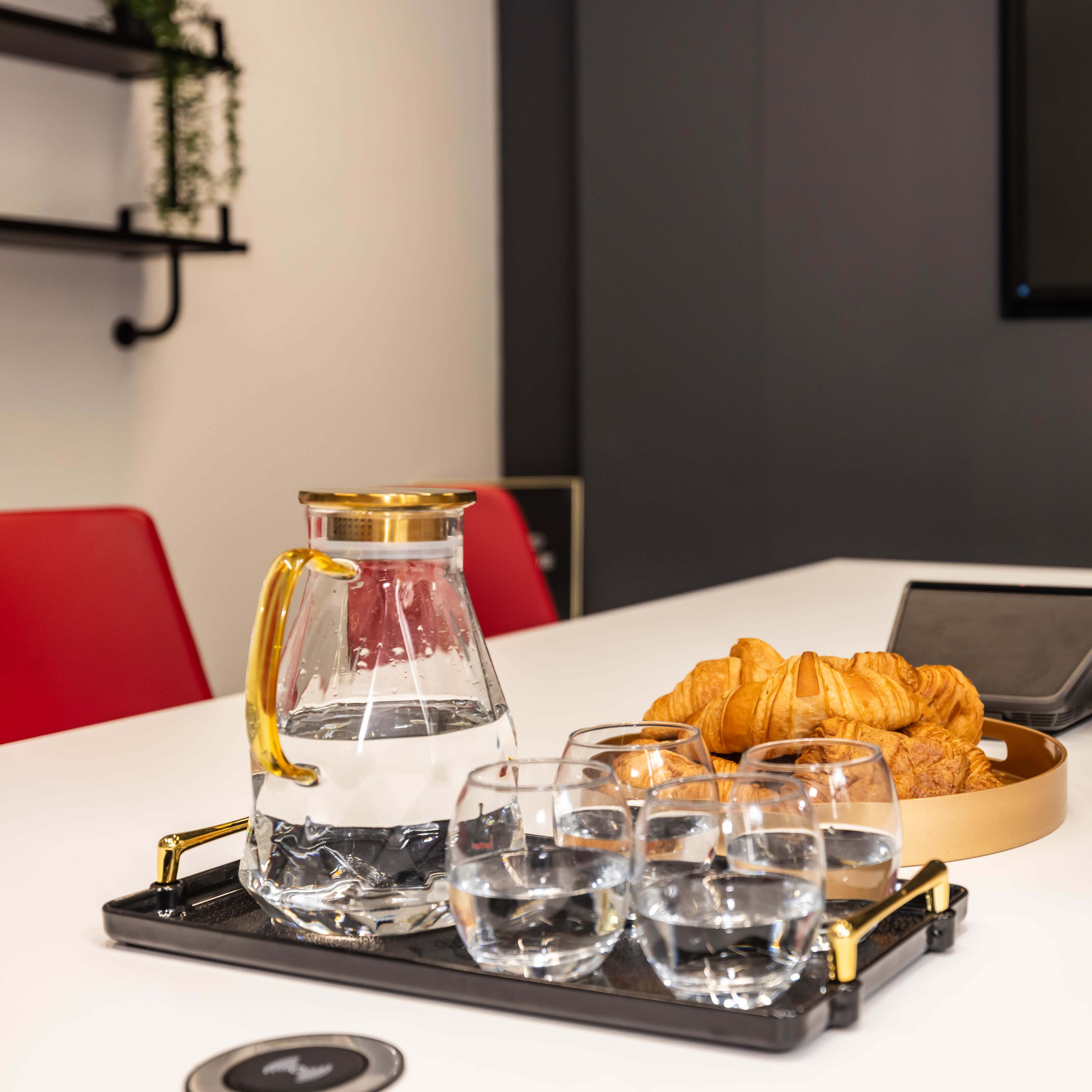 A tray with a glass pitcher of water and several glasses sits beside a plate of croissants on a conference table with red chairs and a wall-mounted screen in the background.