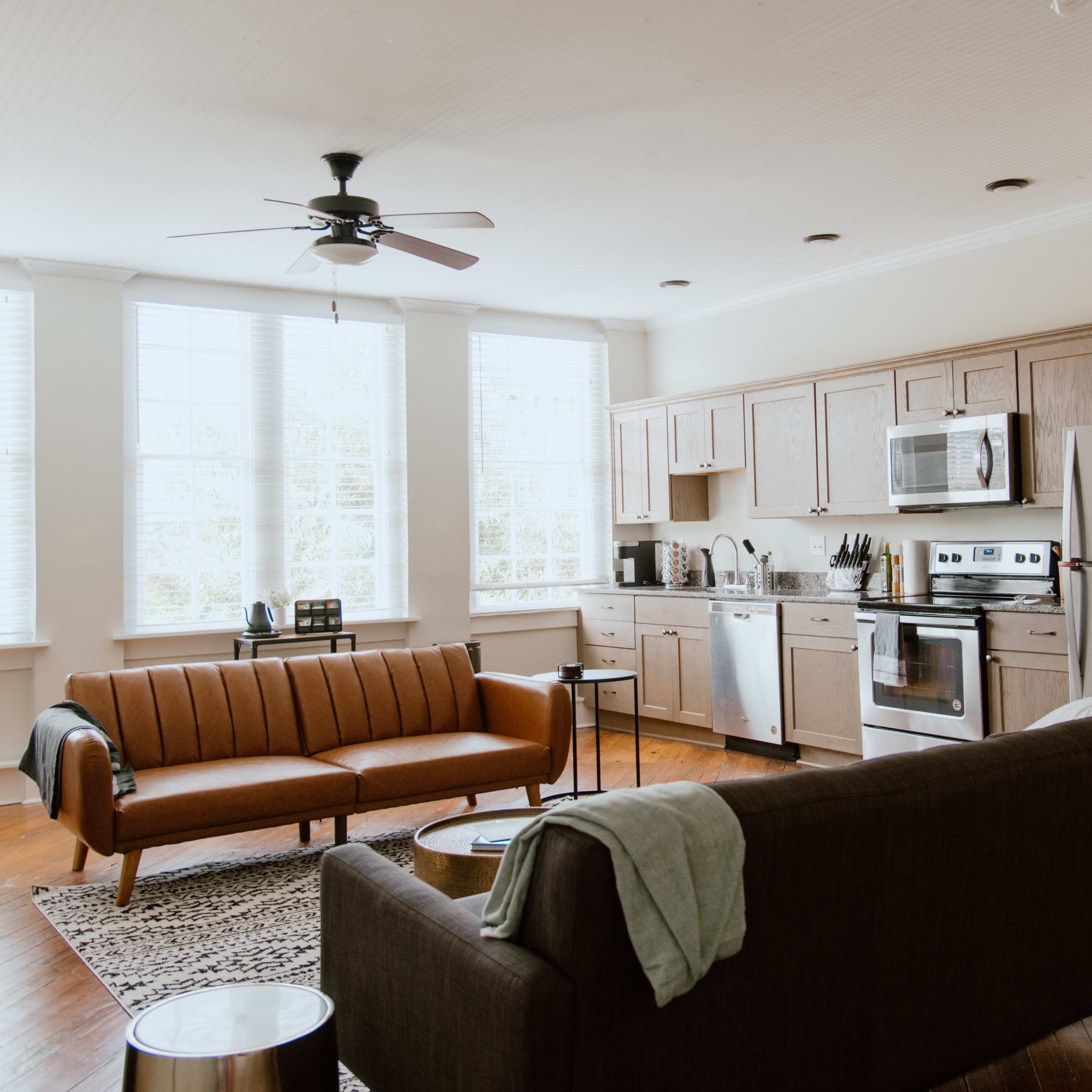 A modern living space featuring a leather sofa, a dark couch, a dining area with a round table, and a kitchen equipped with stainless steel appliances and wooden cabinets.