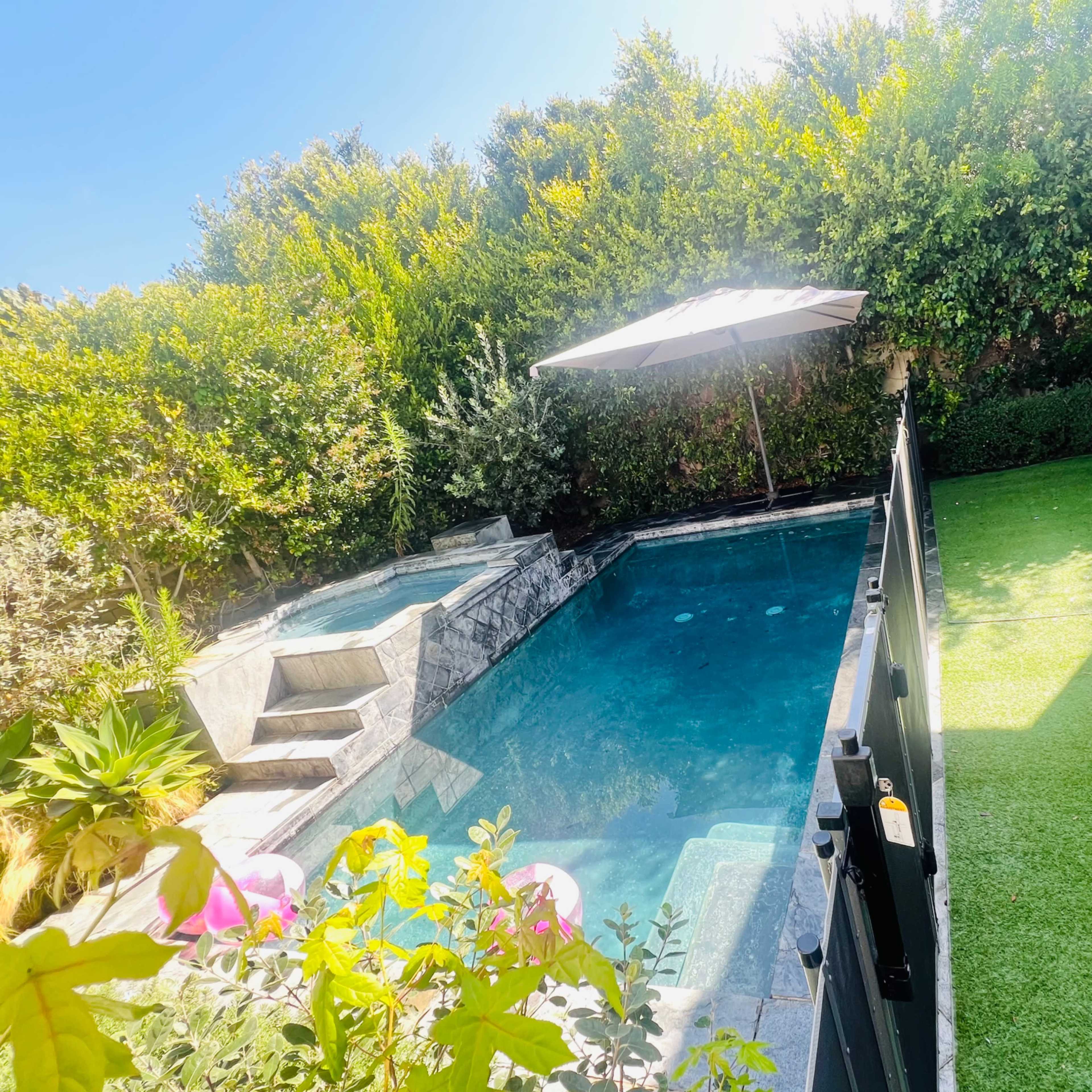 A fenced backyard with a swimming pool, a hot tub, and a shaded seating area under an umbrella, surrounded by lush greenery.