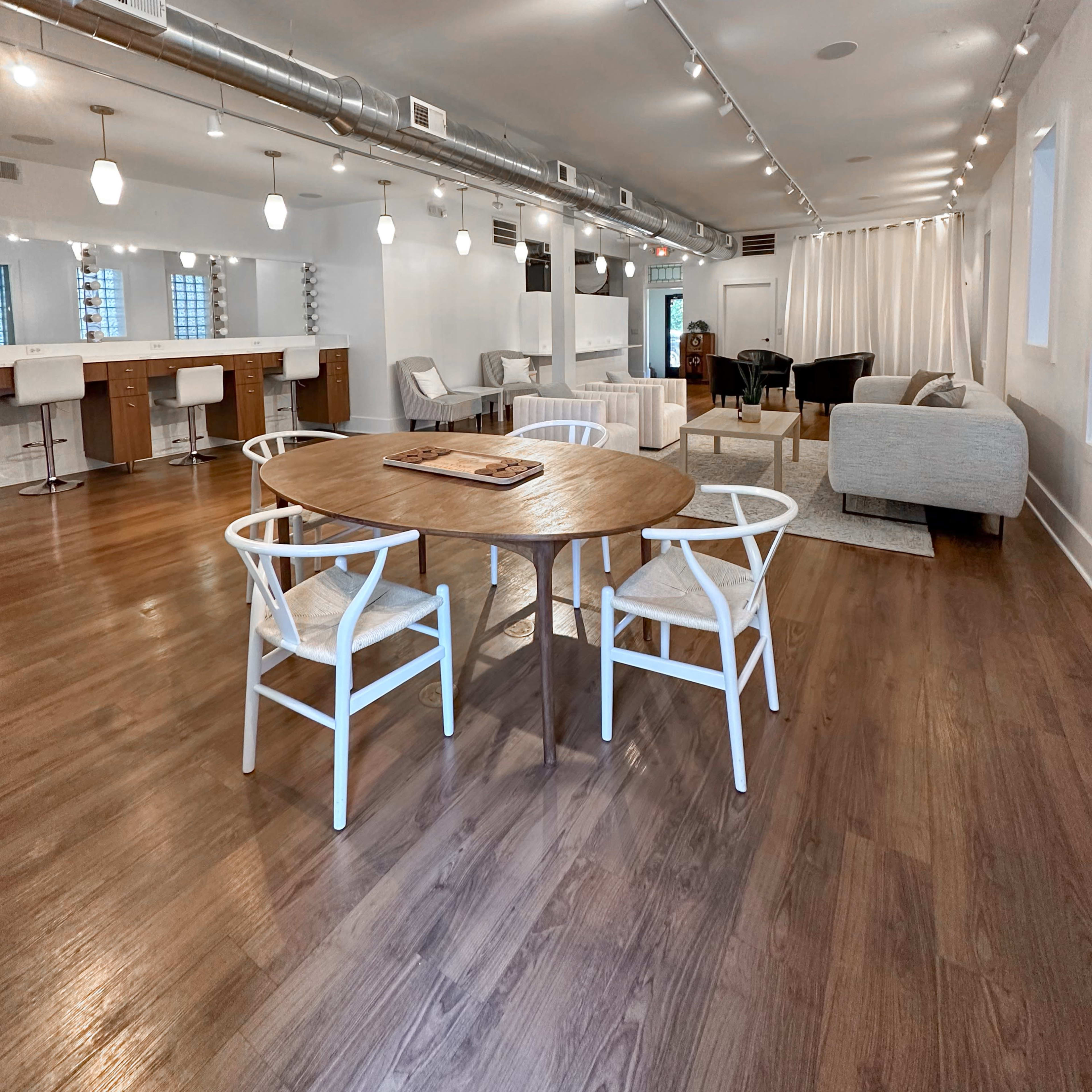 A modern, open-concept interior space featuring a round wooden dining table with white chairs, a seating area with a gray couch, and a bar counter with stools in the background.
