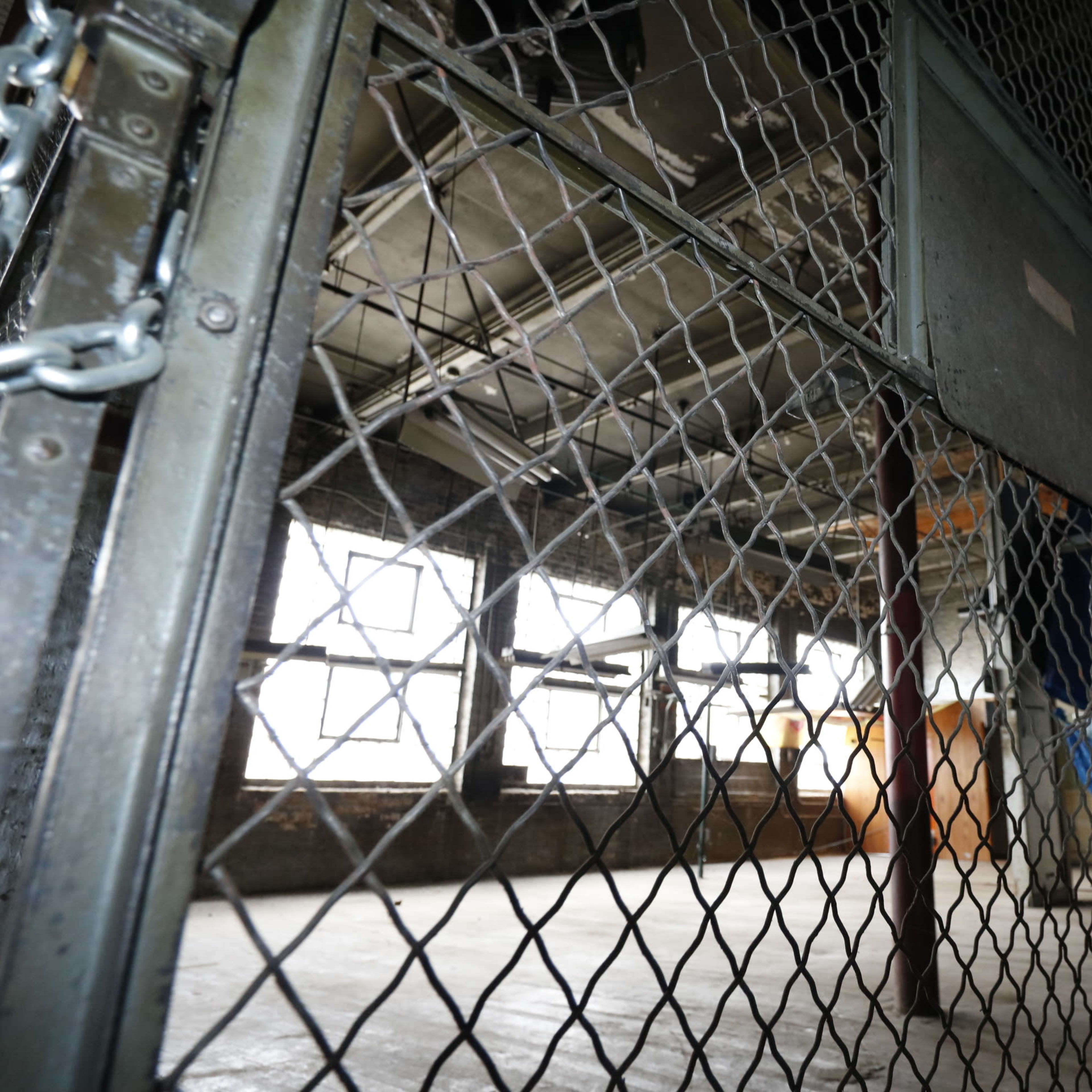 A dimly lit industrial space enclosed by a metal gate with a mesh fence and large windows in the background.