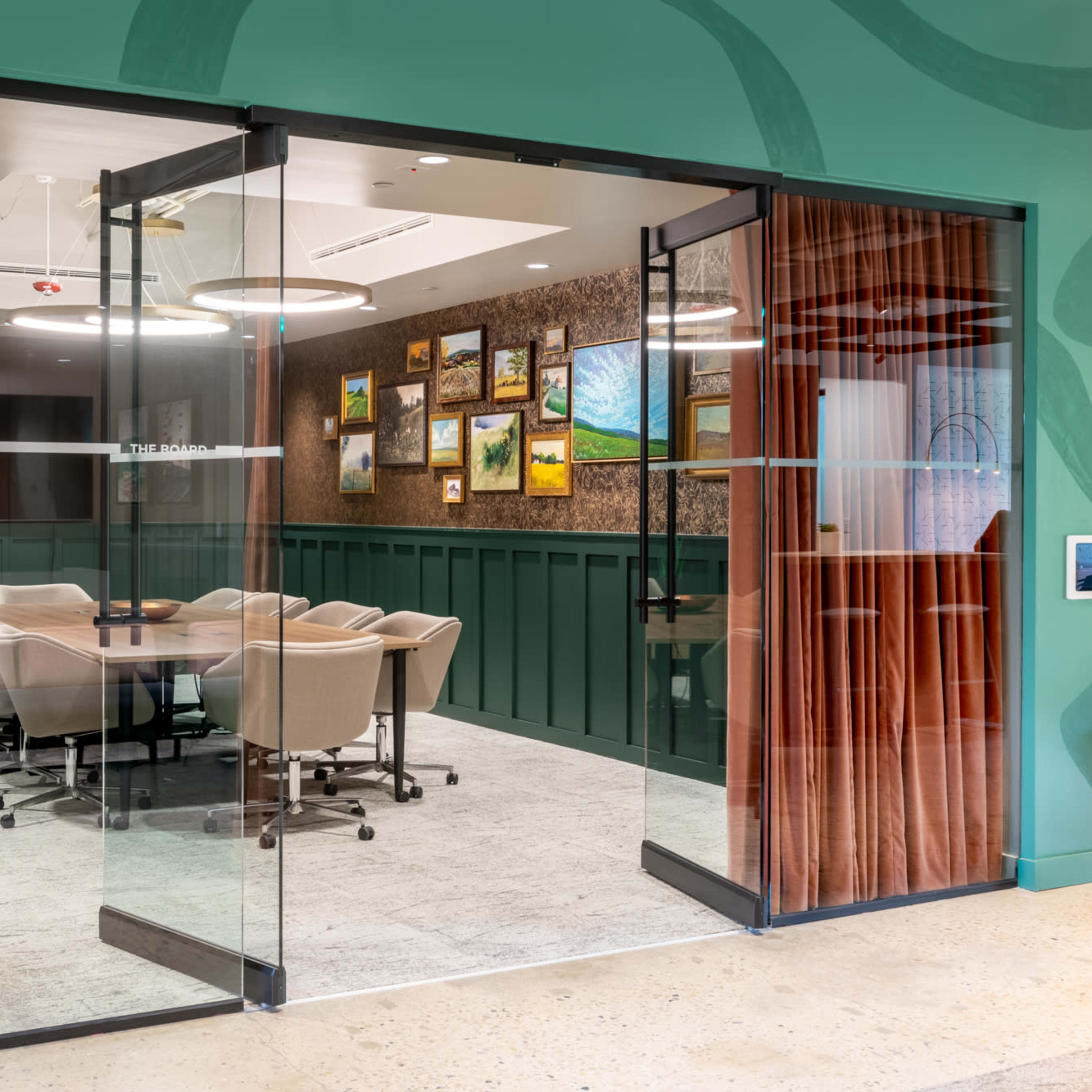 A glass-walled meeting room with a round table and chairs, surrounded by green walls and artwork.