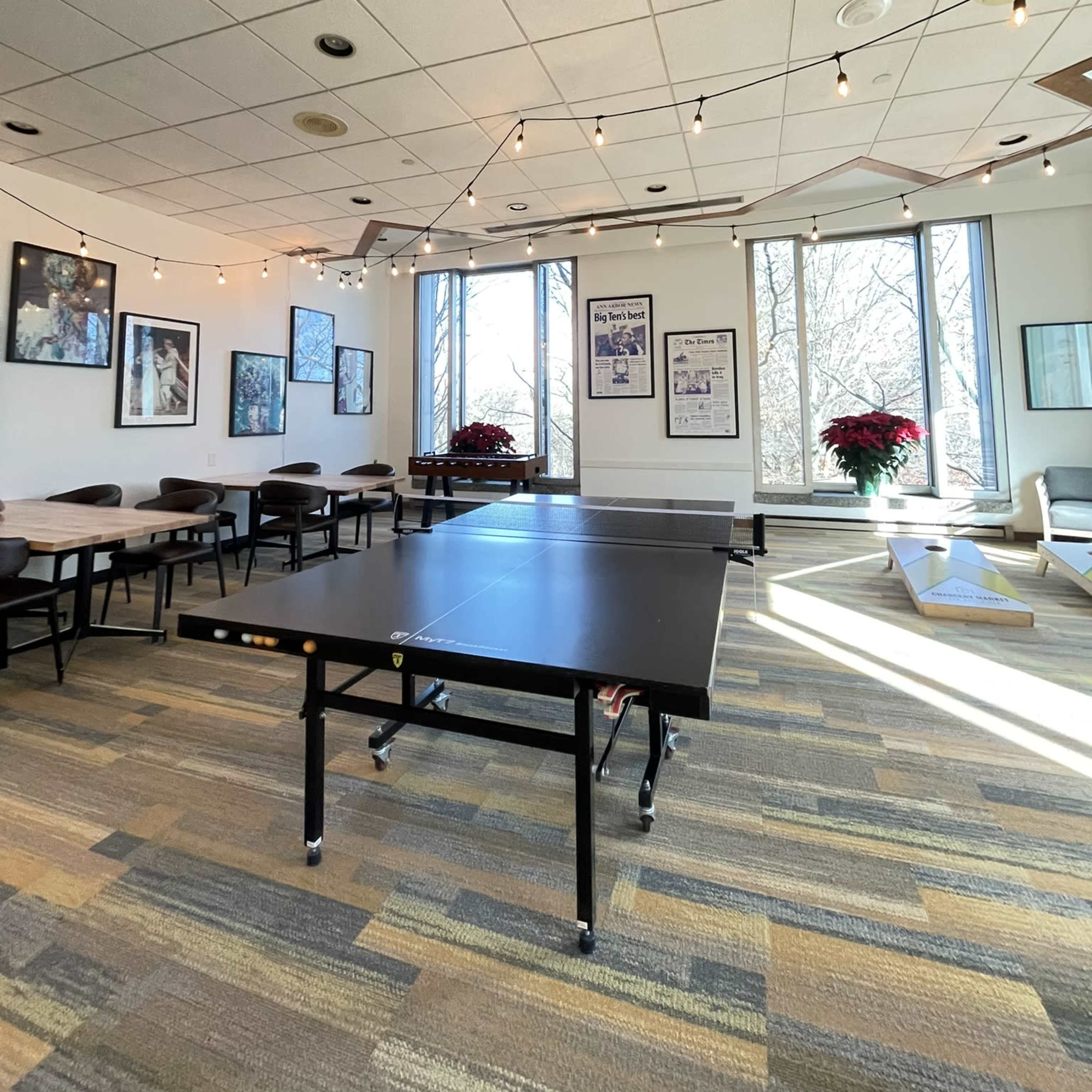 A spacious room features a ping pong table in the center, surrounded by tables and chairs, with large windows allowing natural light to illuminate the space.