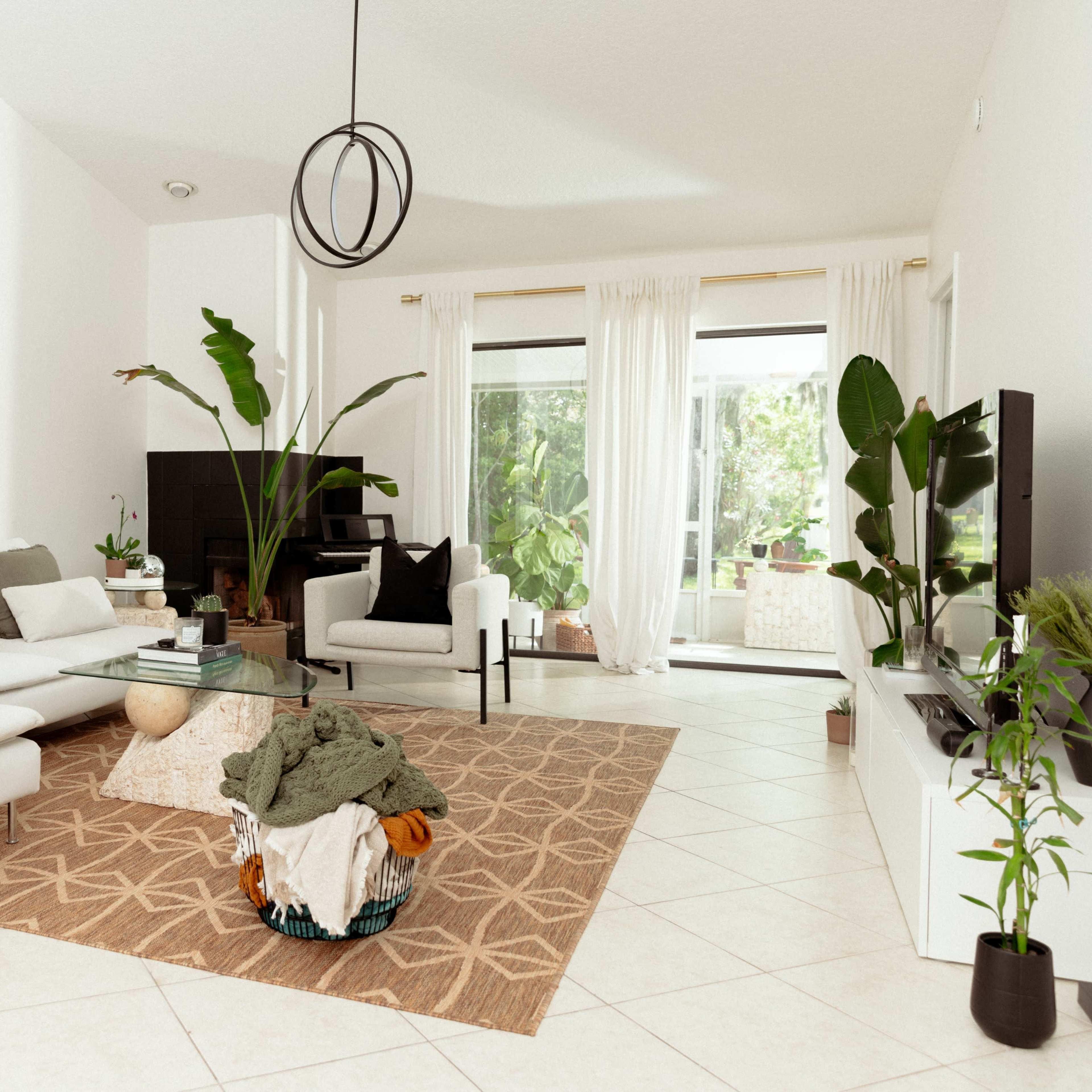The living room features a white sectional sofa with a patterned rug, a coffee table, and large windows that let in natural light, surrounded by indoor plants.