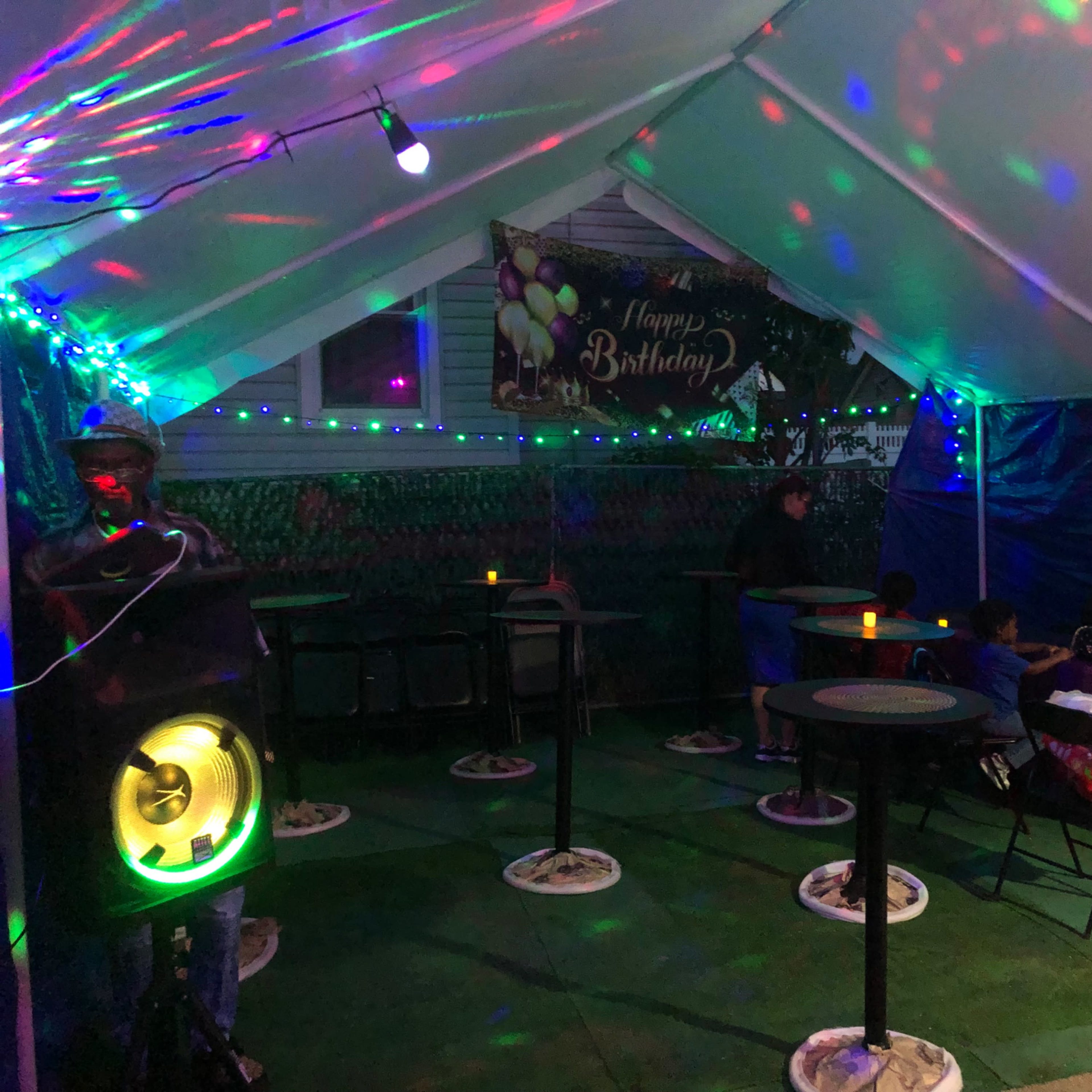A small outdoor tent decorated with a "Happy Birthday" banner has dim lighting and colorful spotlights, with several people seated around tables.