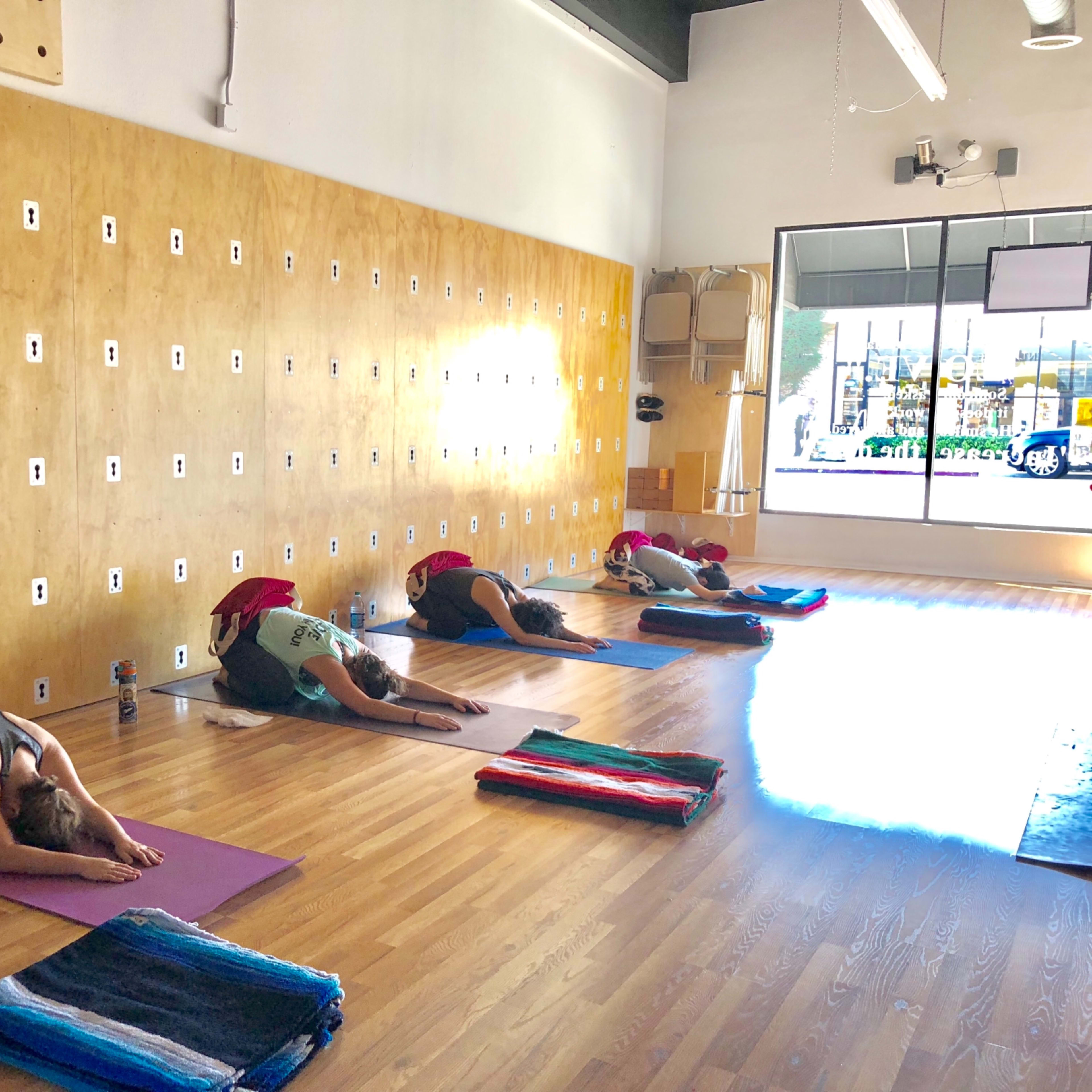 A yoga studio with several individuals practicing a pose on mats, positioned in a row along a wooden floor, against a wall with various hooks.