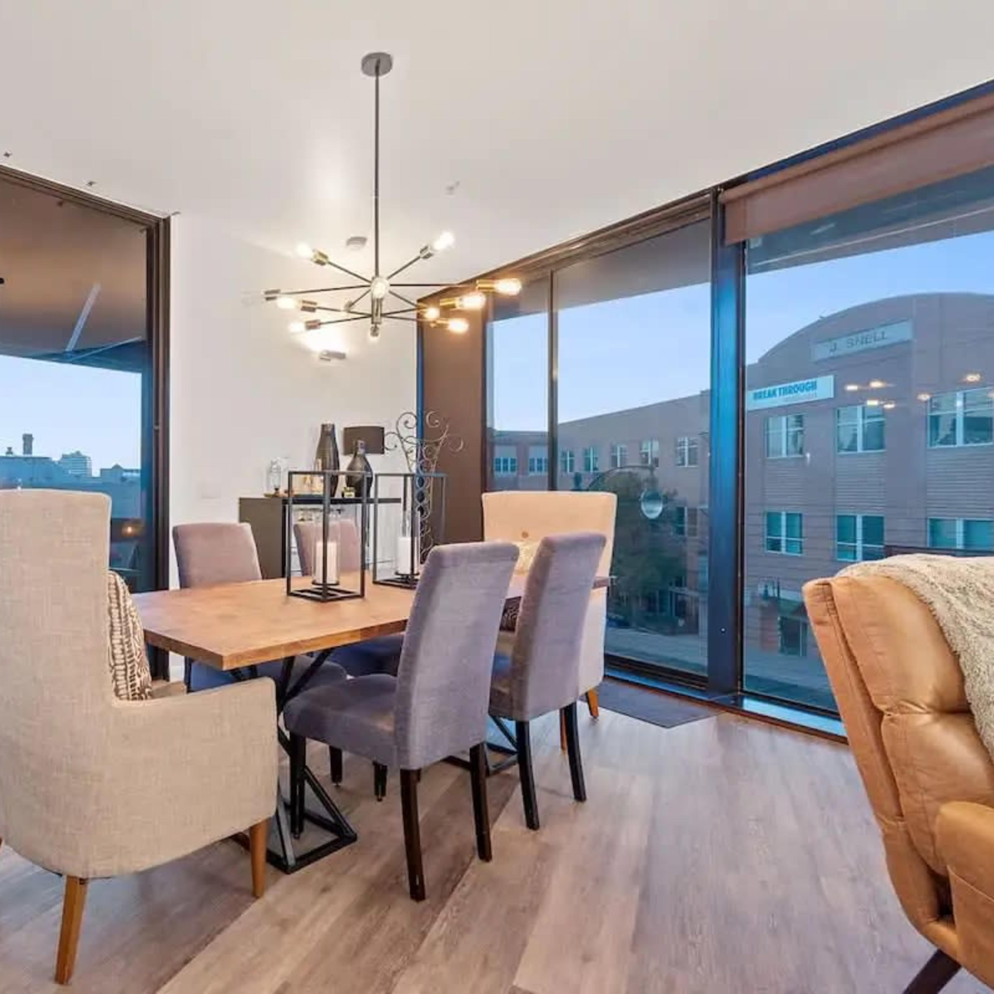 A dining area features a wooden table surrounded by six upholstered chairs, with large glass windows showcasing a balcony view.