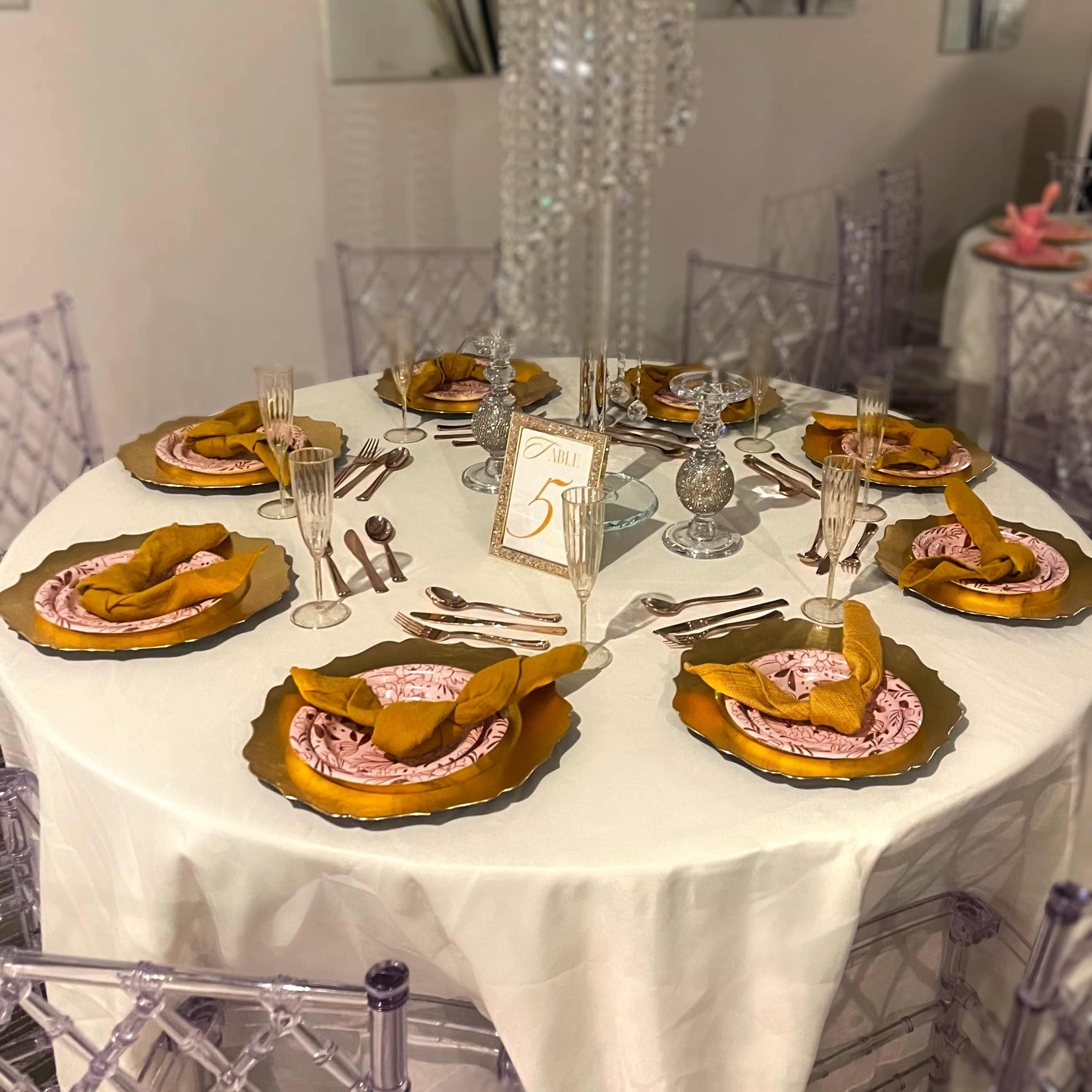 A round dining table is set with elegant plates, silverware, and woven napkins, decorated with a flower centerpiece and a small table number.