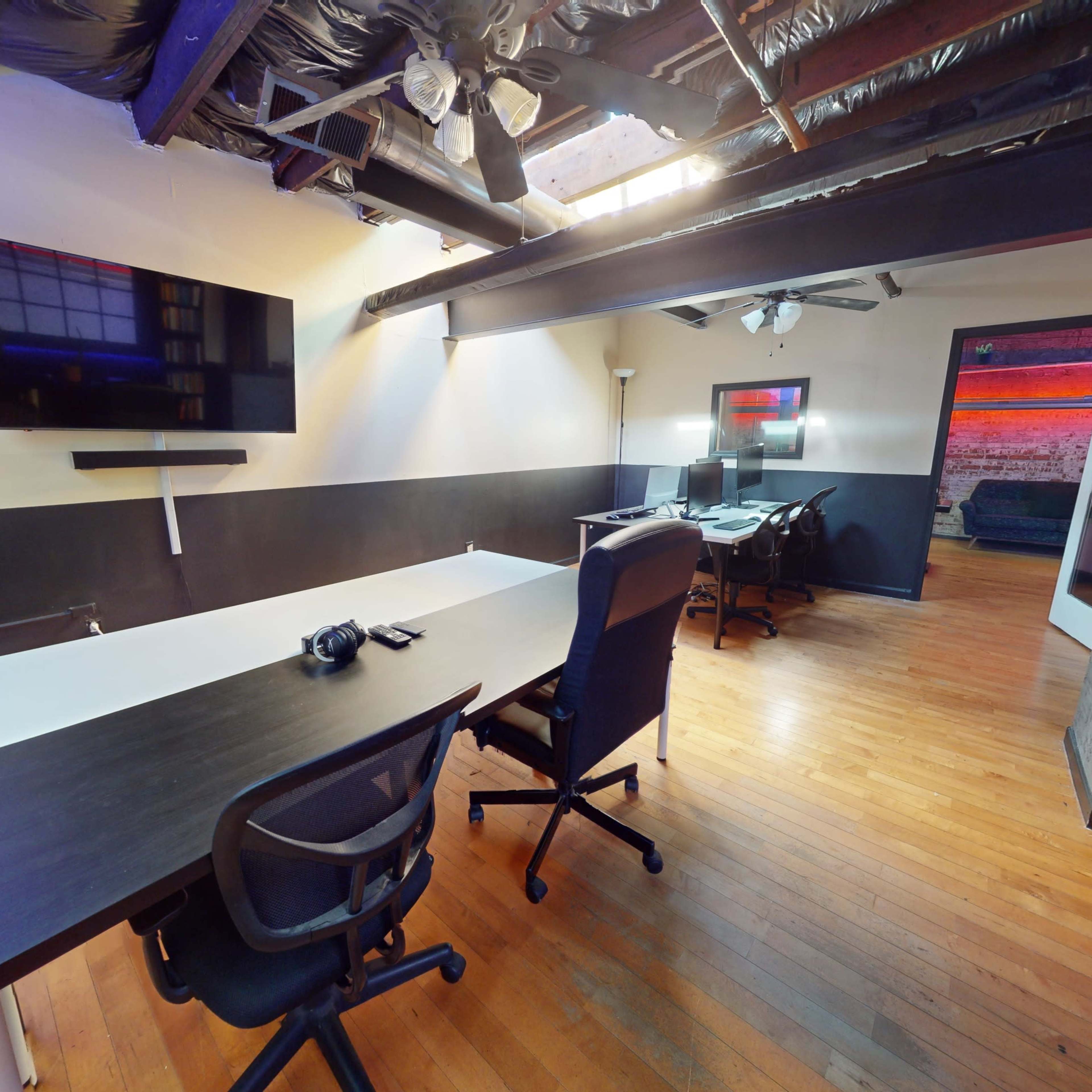 A modern office space with wooden flooring, a large table, several office chairs, and a wall-mounted television, illuminated by natural light from a skylight.