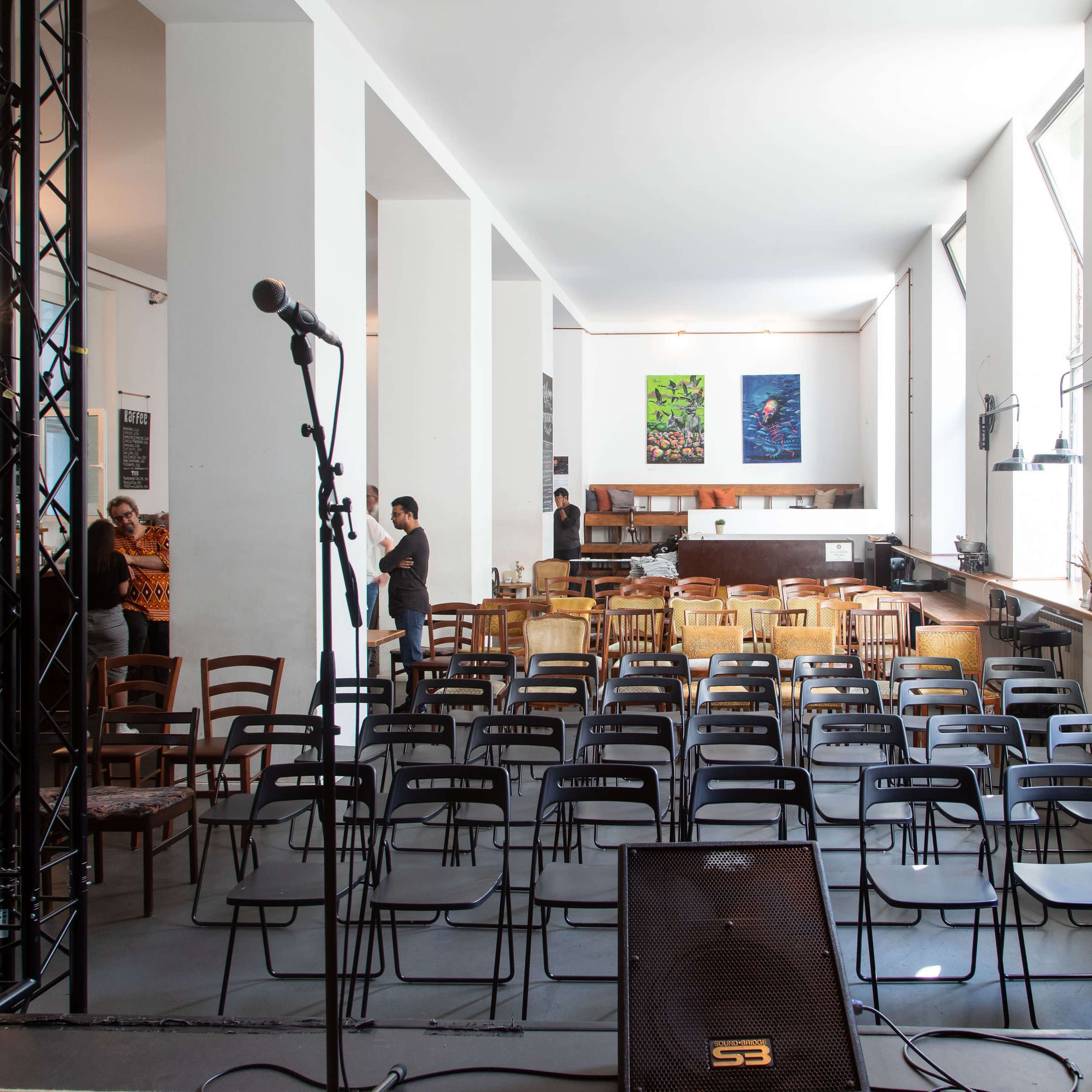 A sparsely furnished performance space with rows of black chairs facing a stage area, flanked by microphones and a bar in the background.