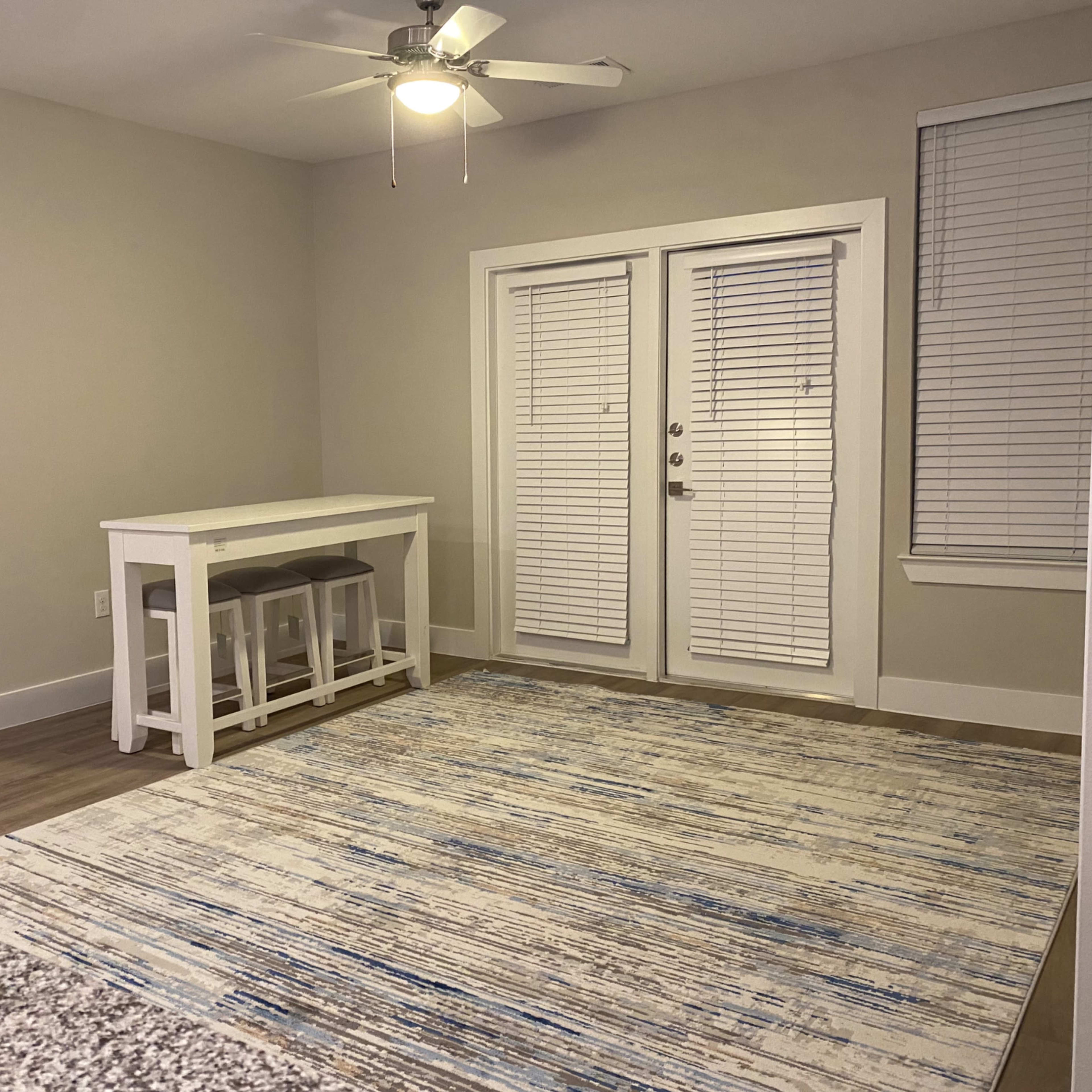 A spacious room featuring a small white dining table with three stools, a large area rug, and double glass doors leading to an outdoor space.