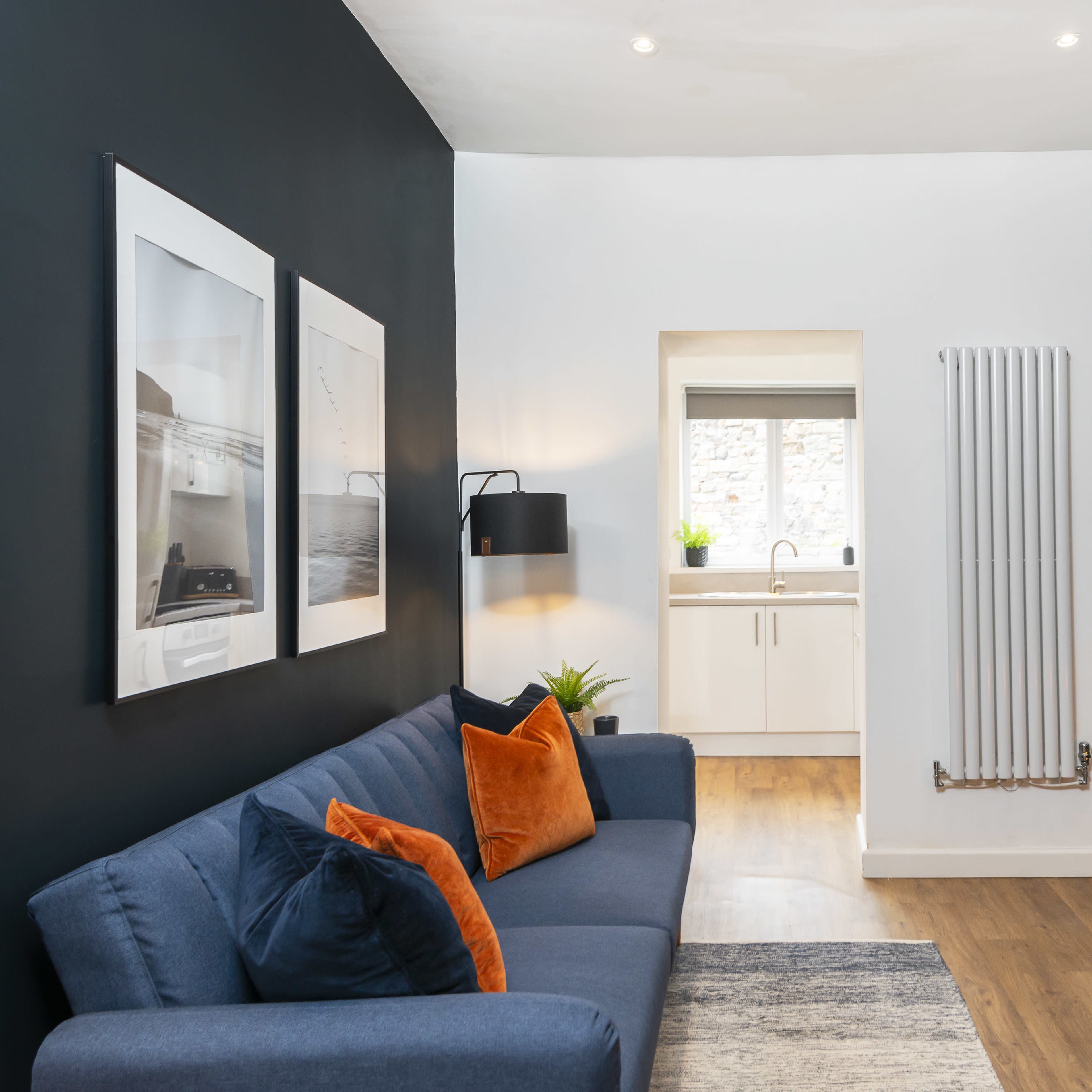 A minimalist living room with a dark accent wall, a blue sofa adorned with orange and blue cushions, and a window leading to a kitchen area.