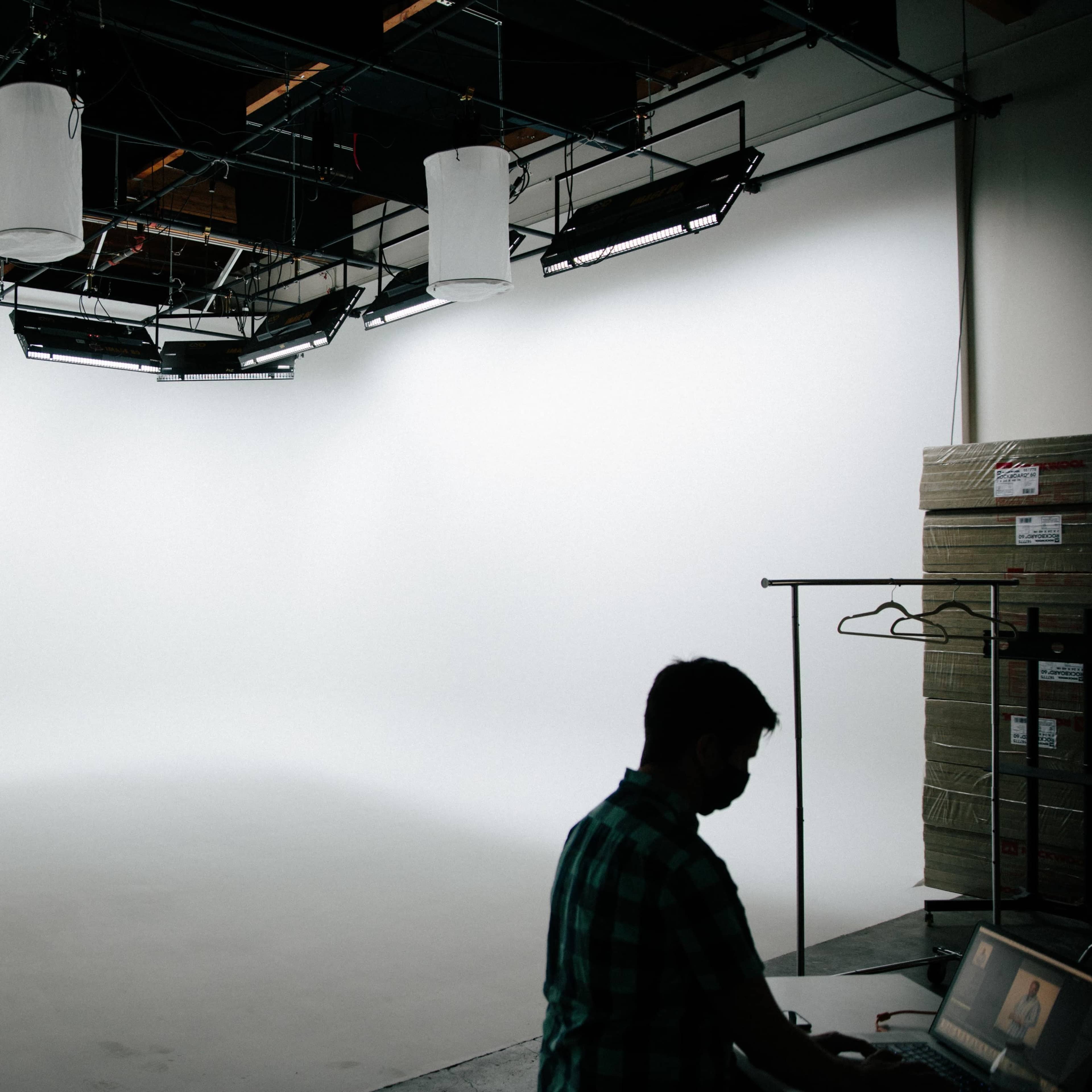 A person works at a table in a spacious, well-lit studio with a seamless white backdrop and overhead lights.