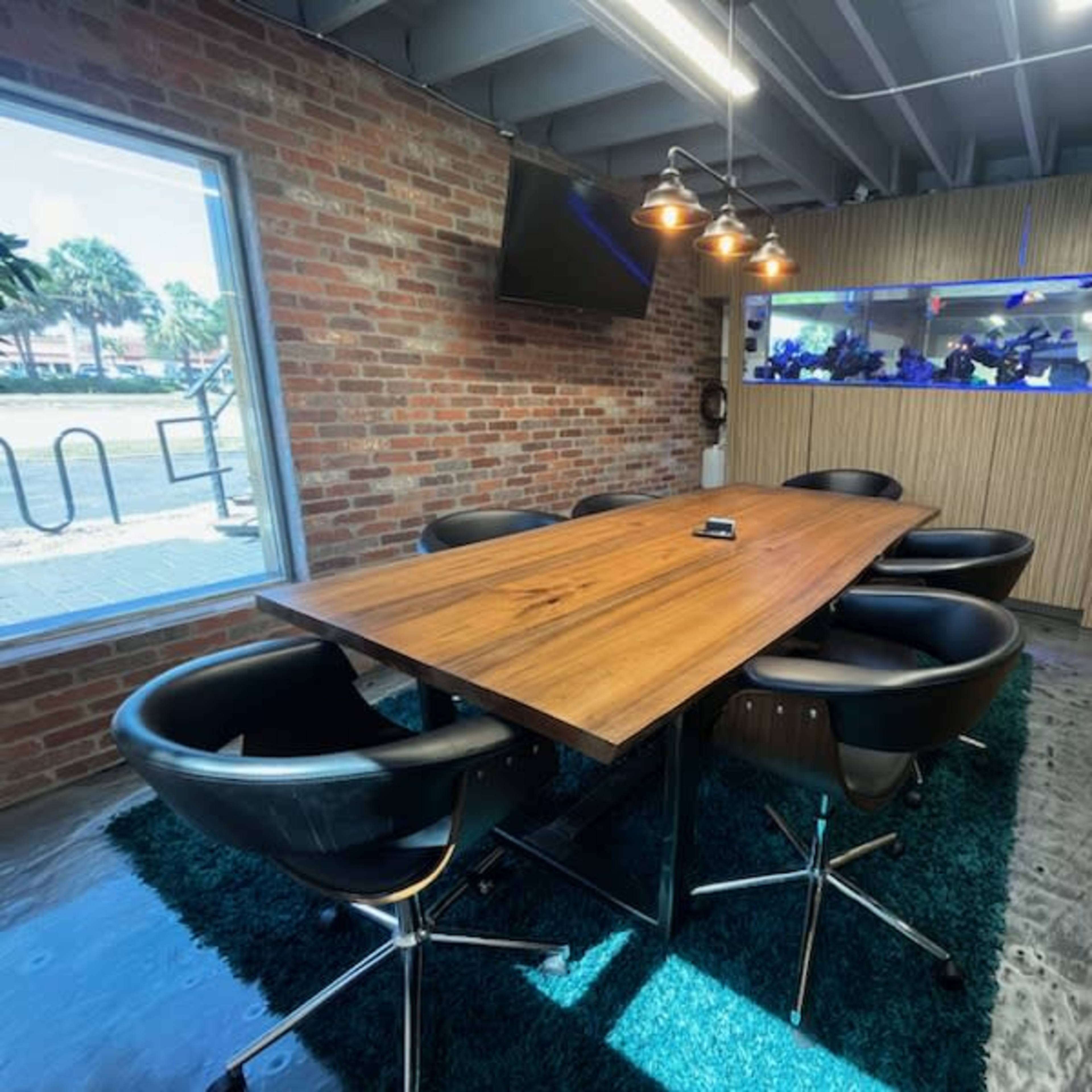 A large wooden conference table surrounded by black chairs is situated in a room with brick walls and a glass partition, featuring a view of an aquarium.