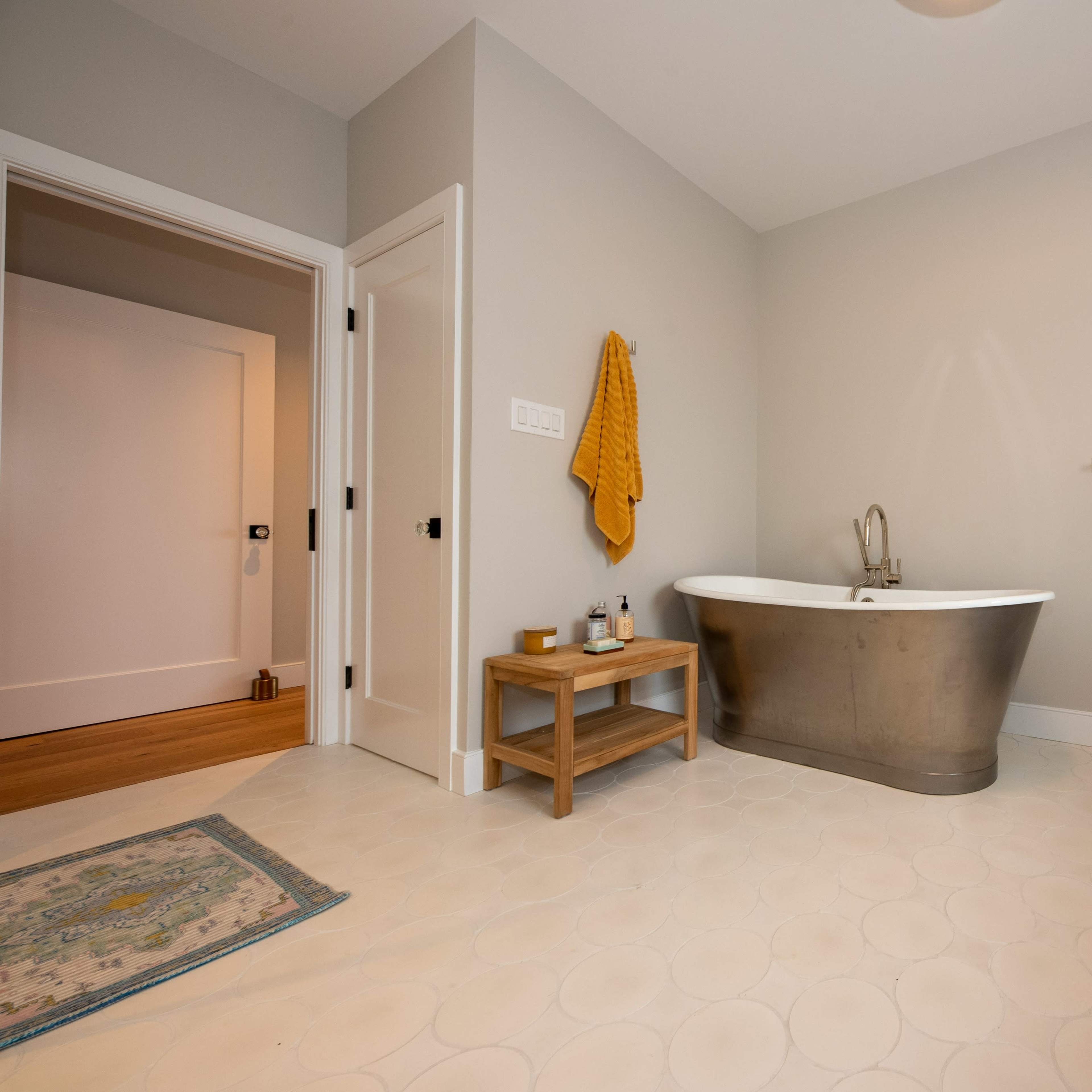A modern bathroom featuring a freestanding metal bathtub, a wooden bench, and light-colored tile flooring.