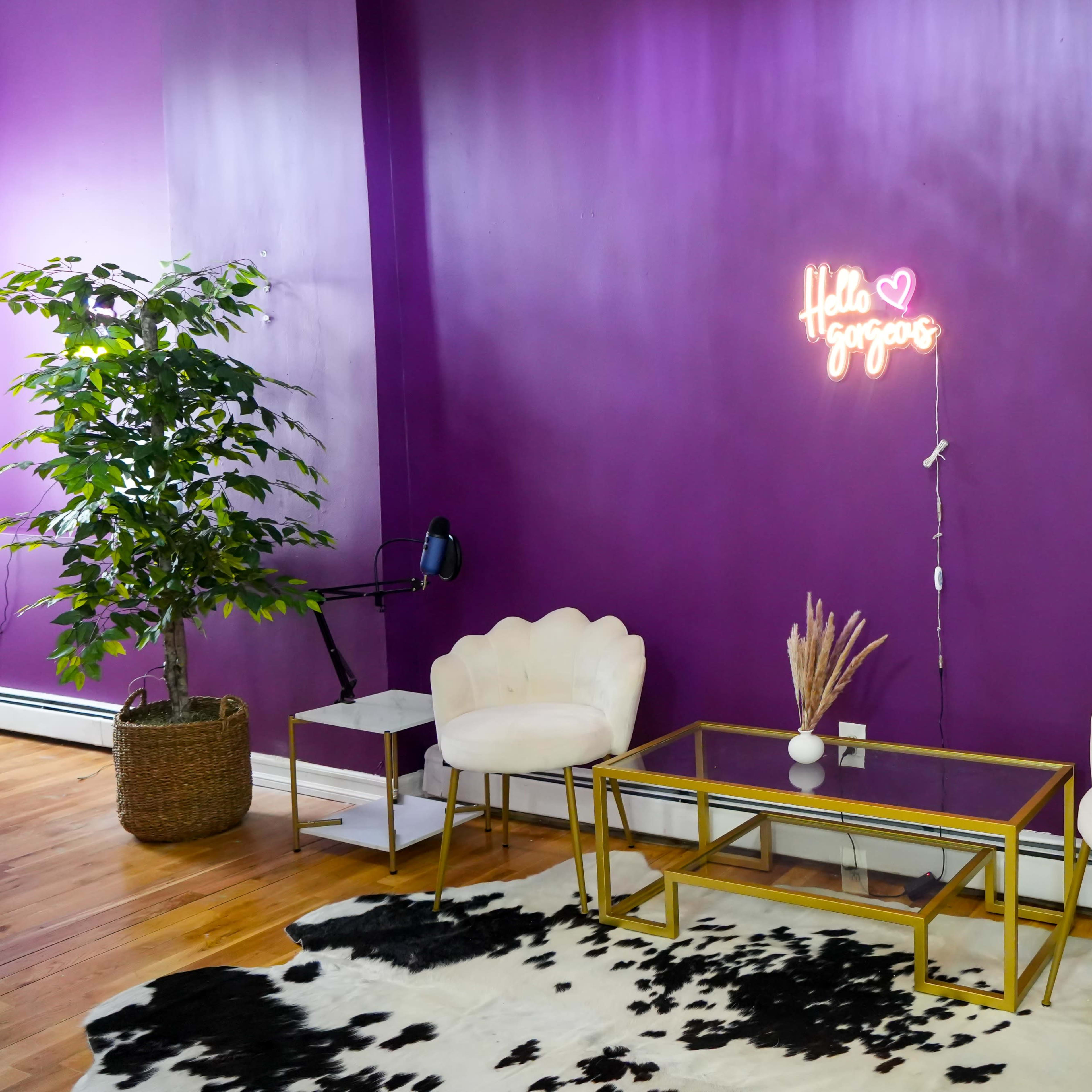 A seating area with two white chairs and a glass table on a black and white cowhide rug, beneath a purple wall featuring a neon sign that says "Hello Gorgeous."