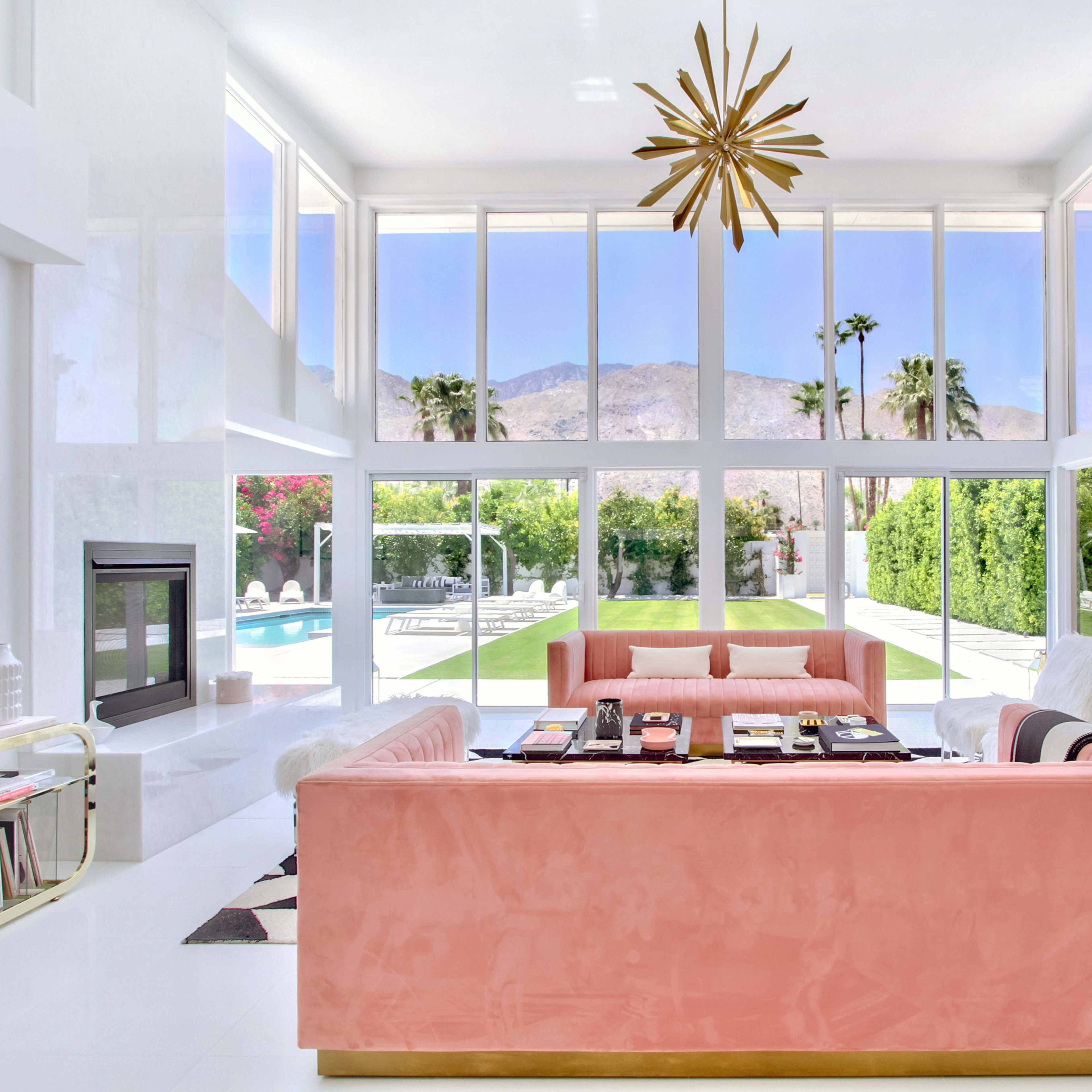 The living room features two large sofas facing a wall of windows that overlook a pool and palm trees with mountains in the background.