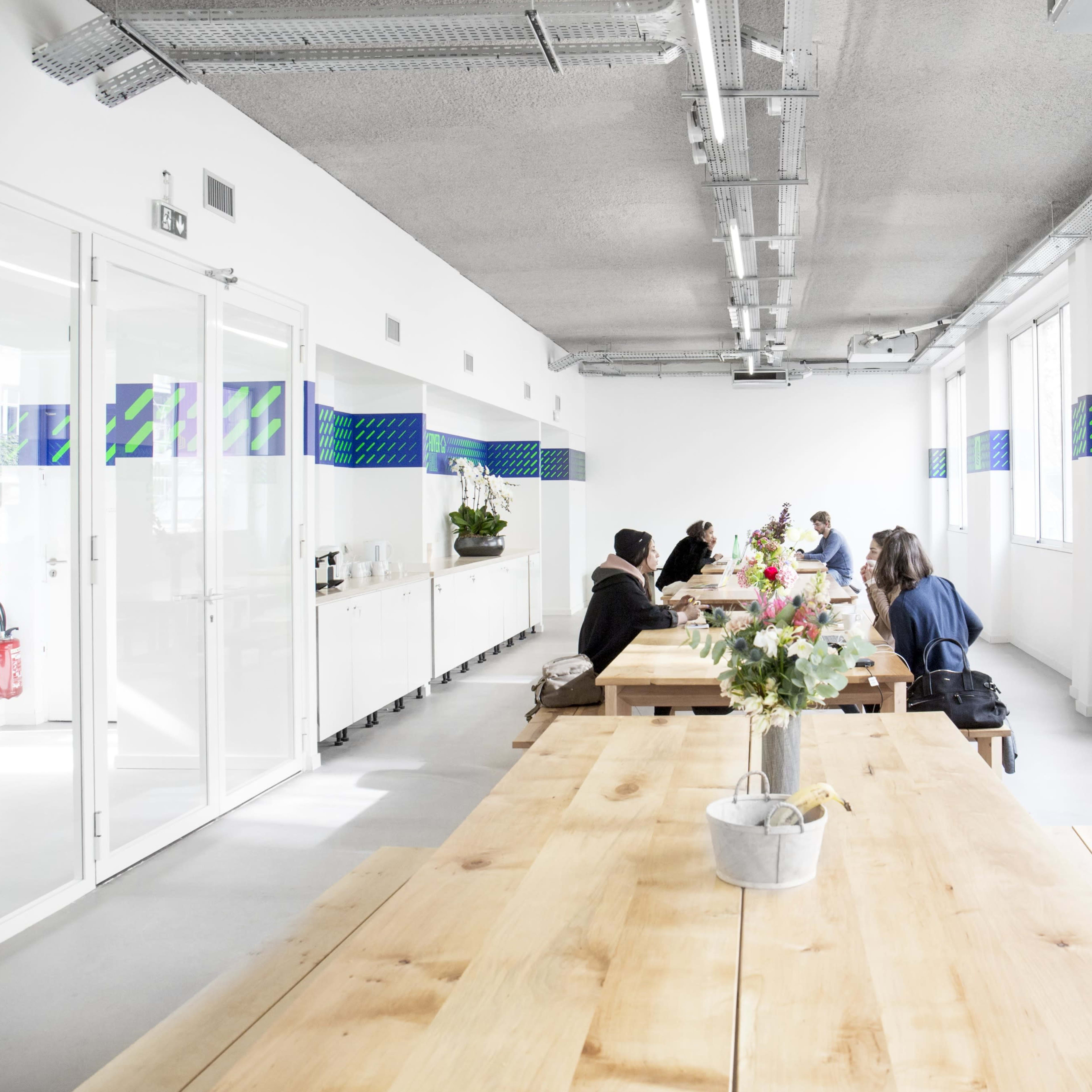 A long wooden table is surrounded by several people in a bright, modern office space with large windows and decorative wall designs.