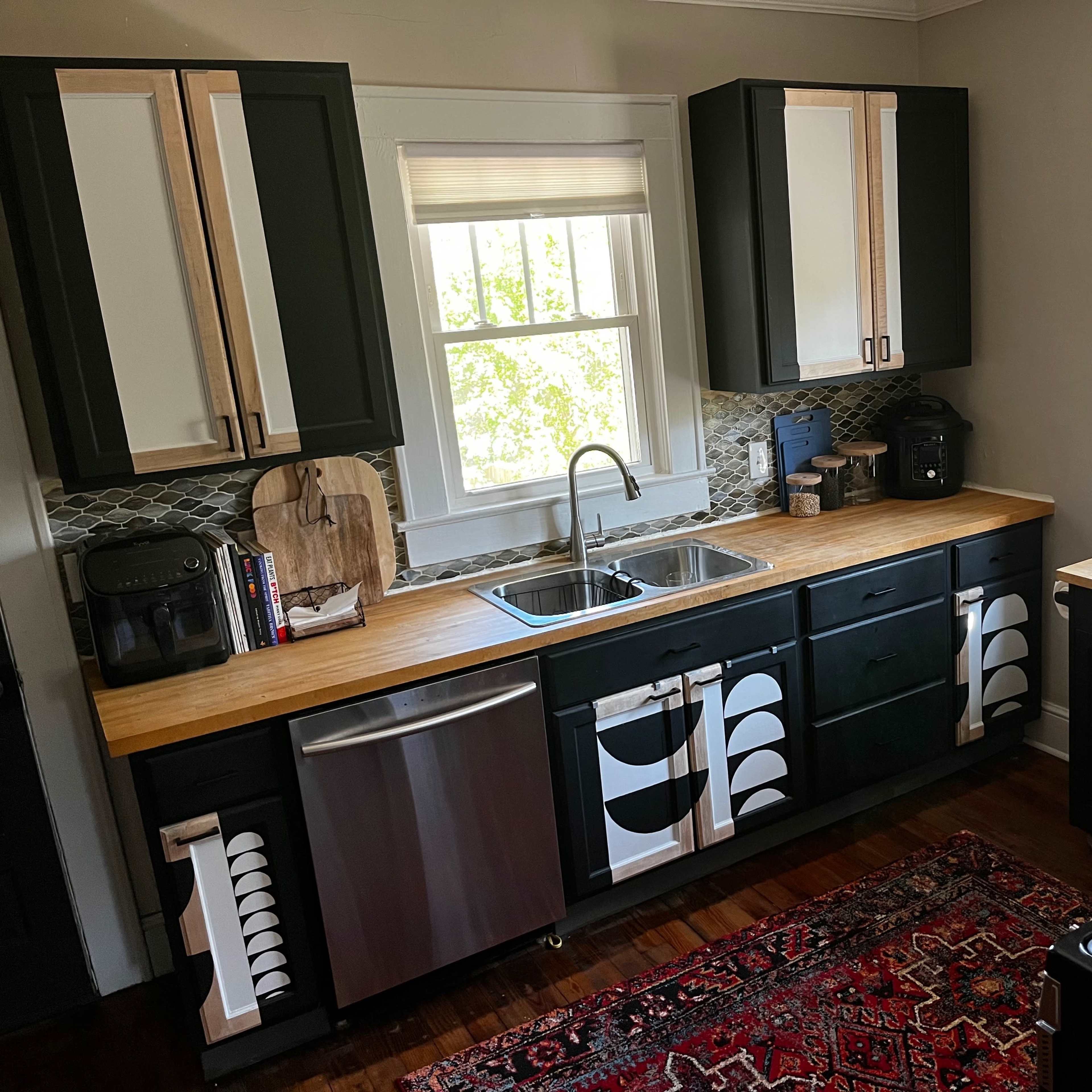 A modern kitchen features black and natural wood cabinetry, a stainless steel sink, and a gas stove with a patterned rug on the wooden floor.