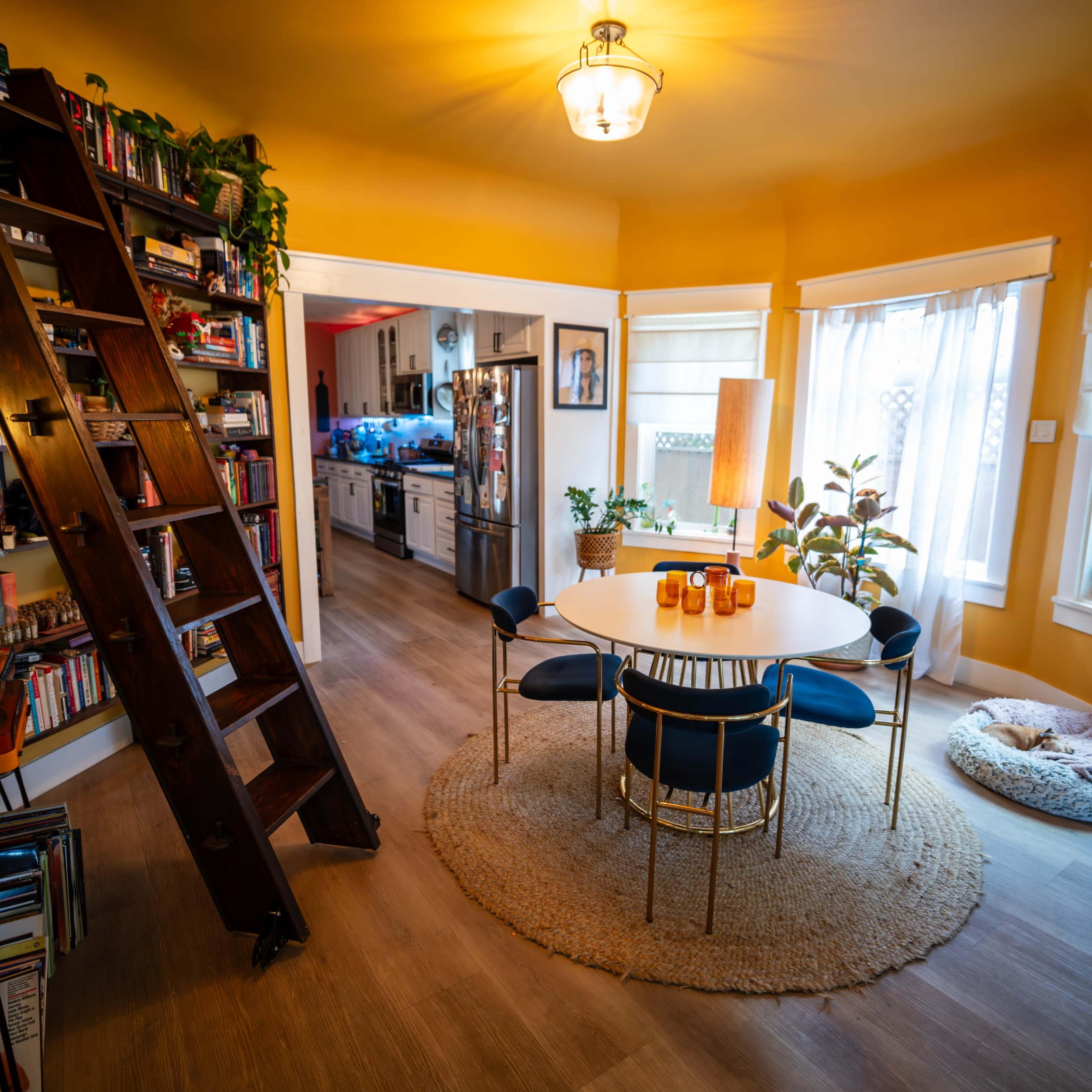 A warm-toned dining area features a round table with blue chairs, surrounded by plants and a wooden ladder leading to an upper shelf, while a kitchen is visible in the background.