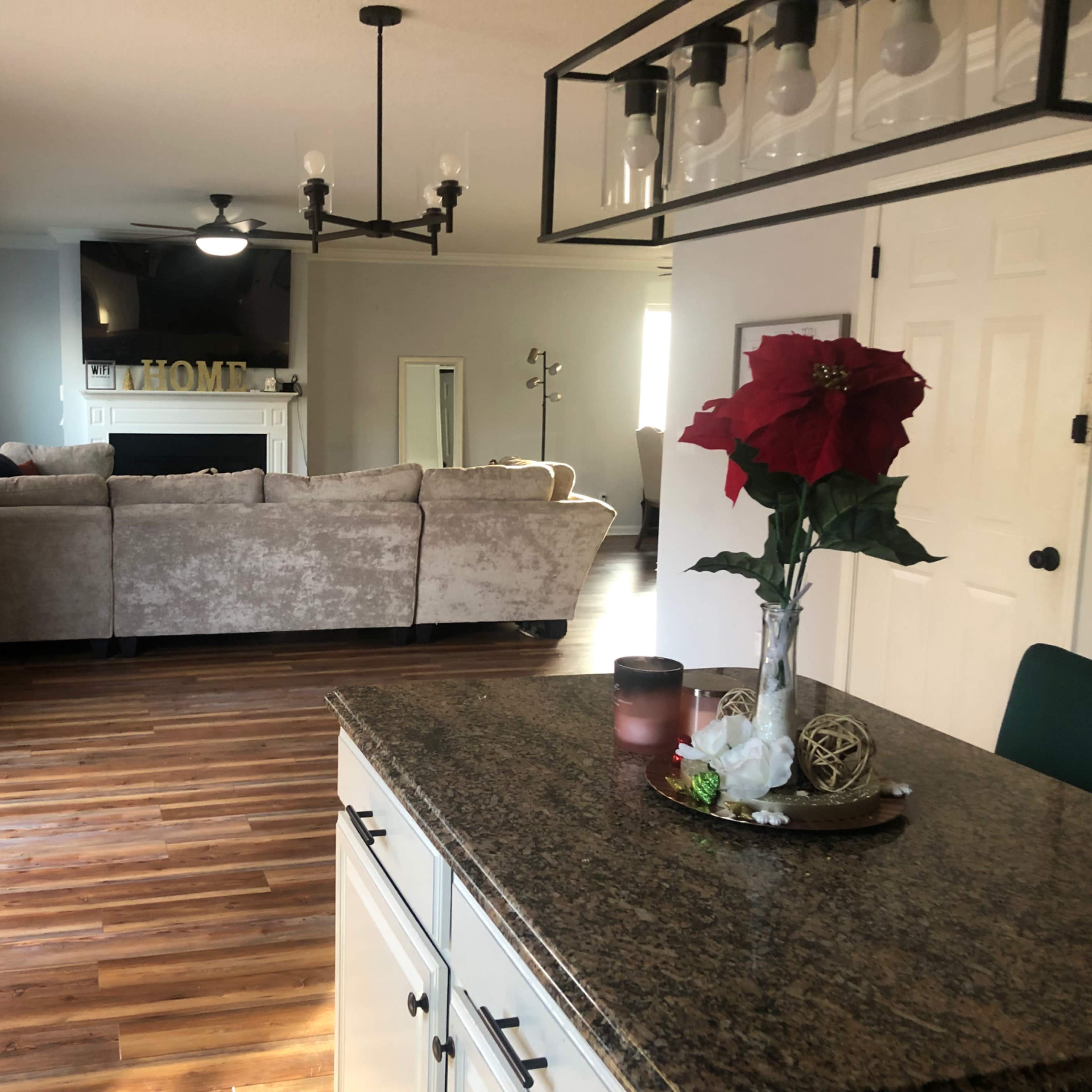 A living room with a sofa, a television mounted above a fireplace, and a kitchen area with a countertop and a decorative floral arrangement.