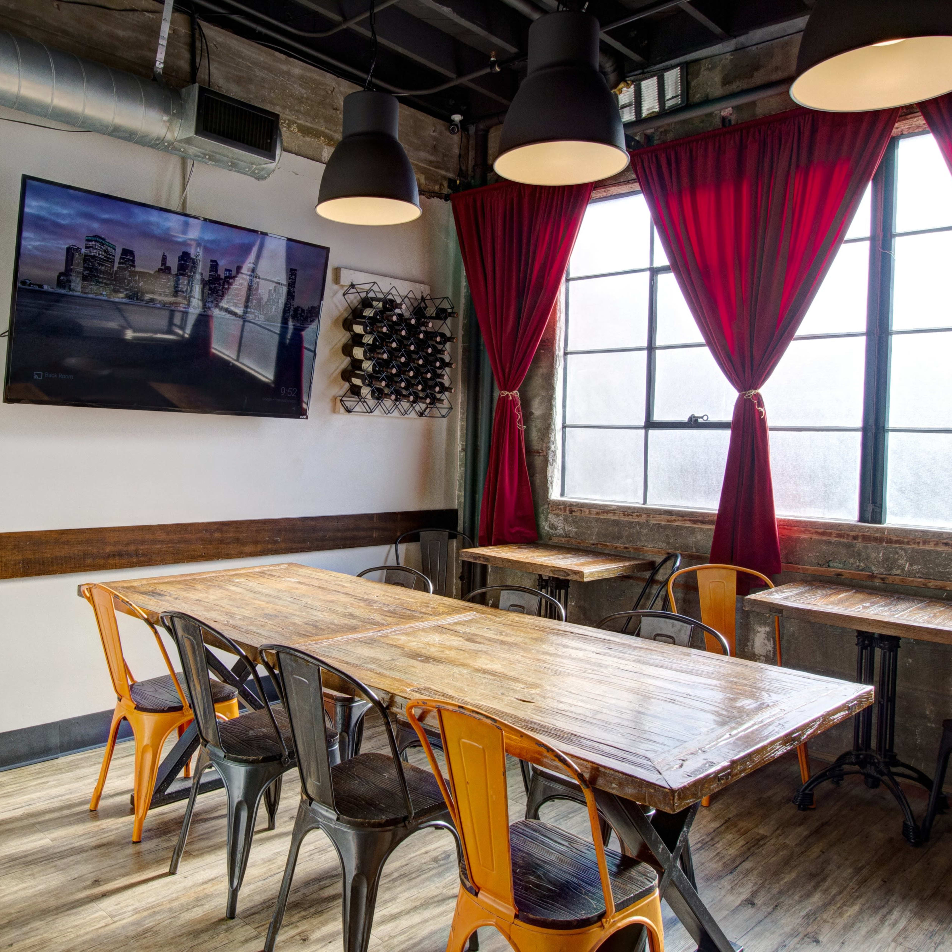 A dining area features a wooden table surrounded by metal chairs, with a large television mounted on the wall and red curtains framing a window.