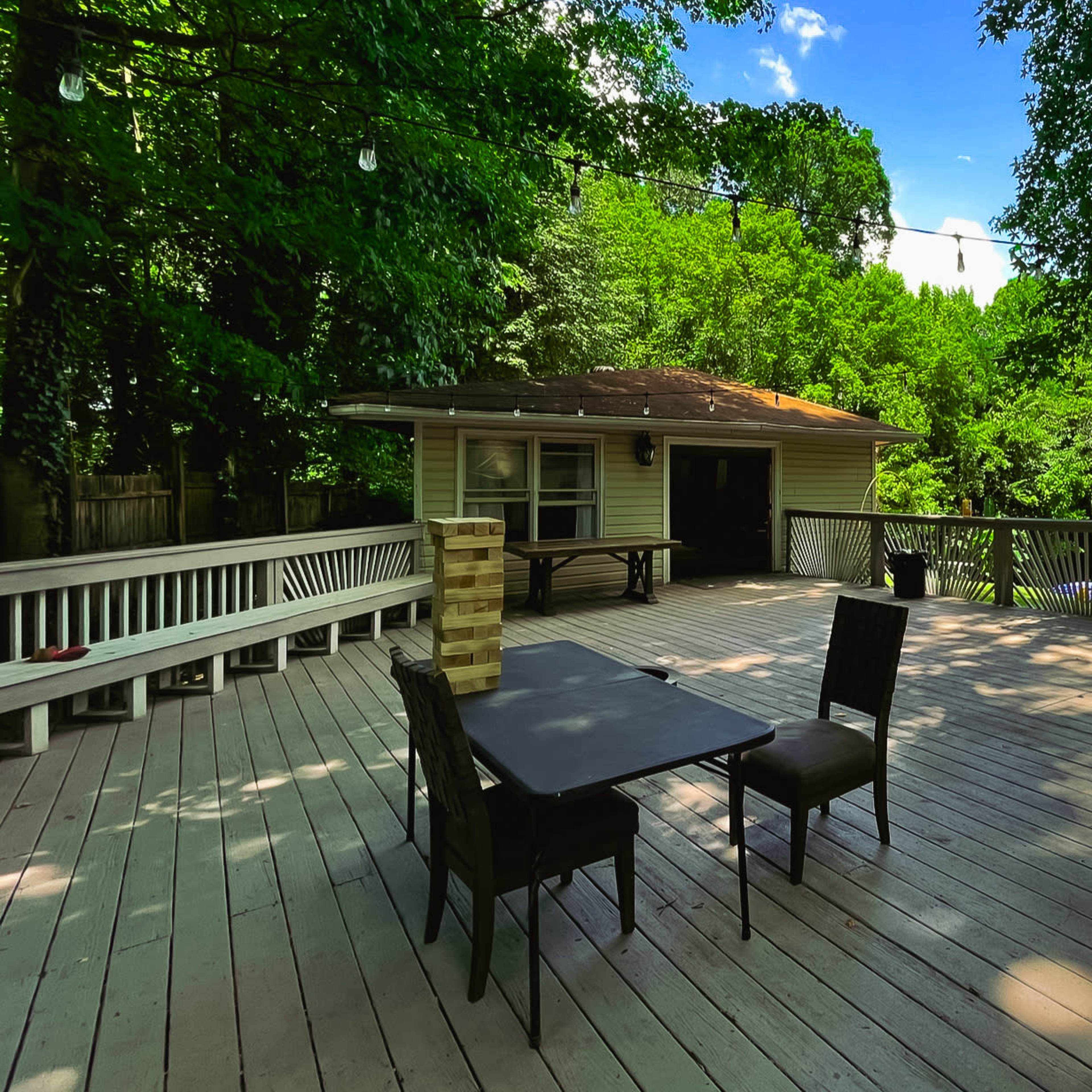 A spacious wooden deck features a table and chairs, surrounded by trees and overlooking a nearby pool and buildings.