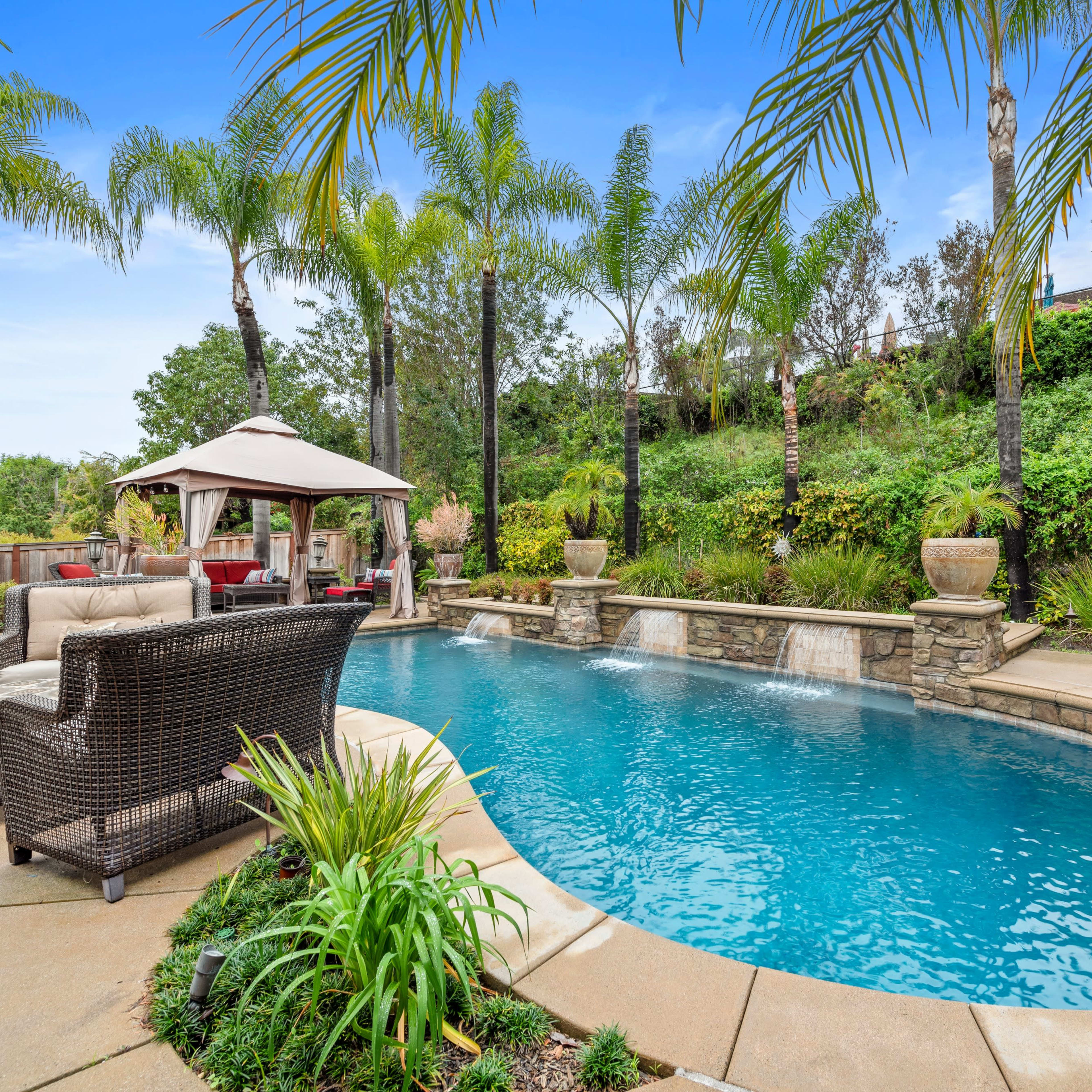 The scene features a curved swimming pool surrounded by palm trees, with a gazebo and seating area on the patio.