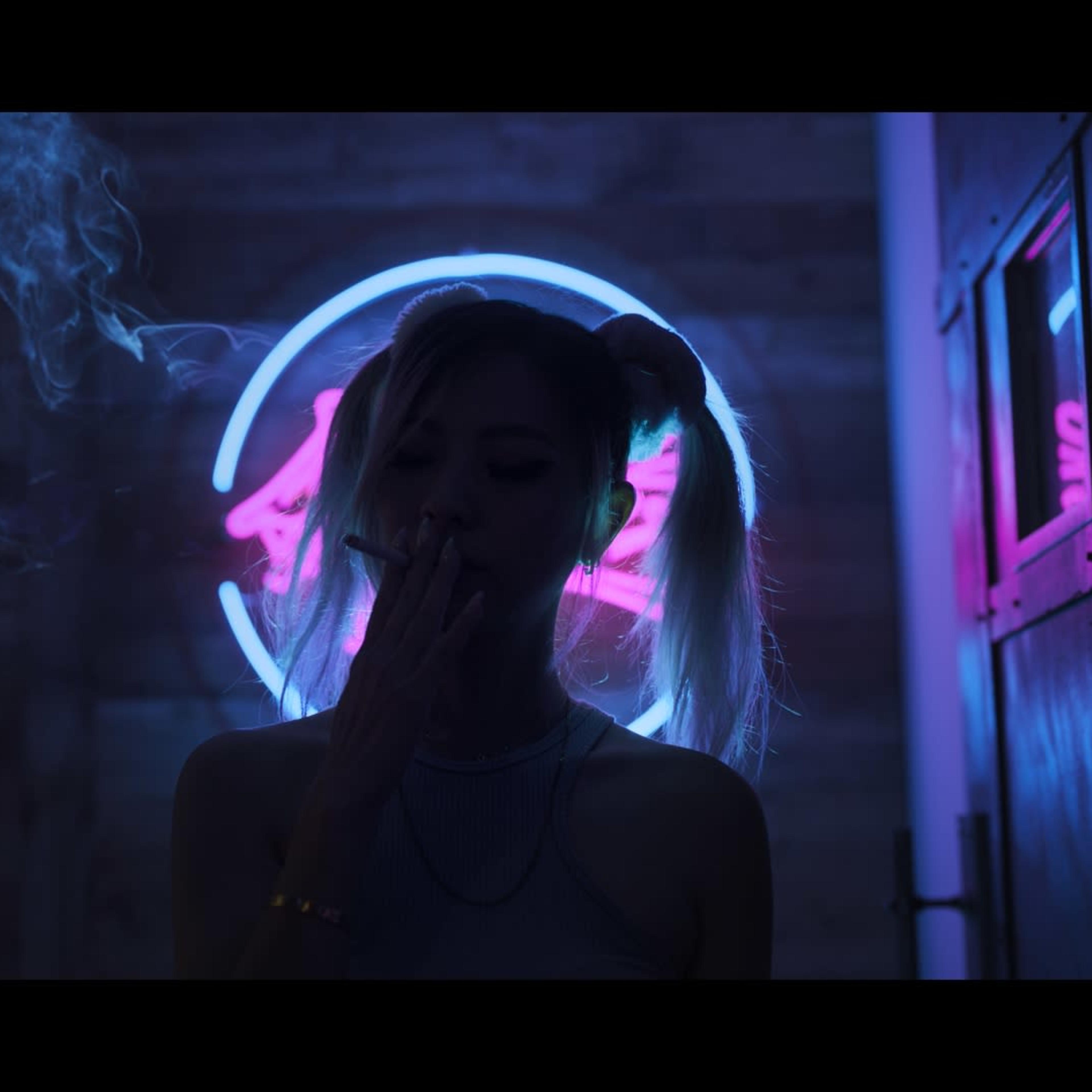 A woman with two ponytails smokes a cigarette in front of a glowing neon sign in a dimly lit setting.