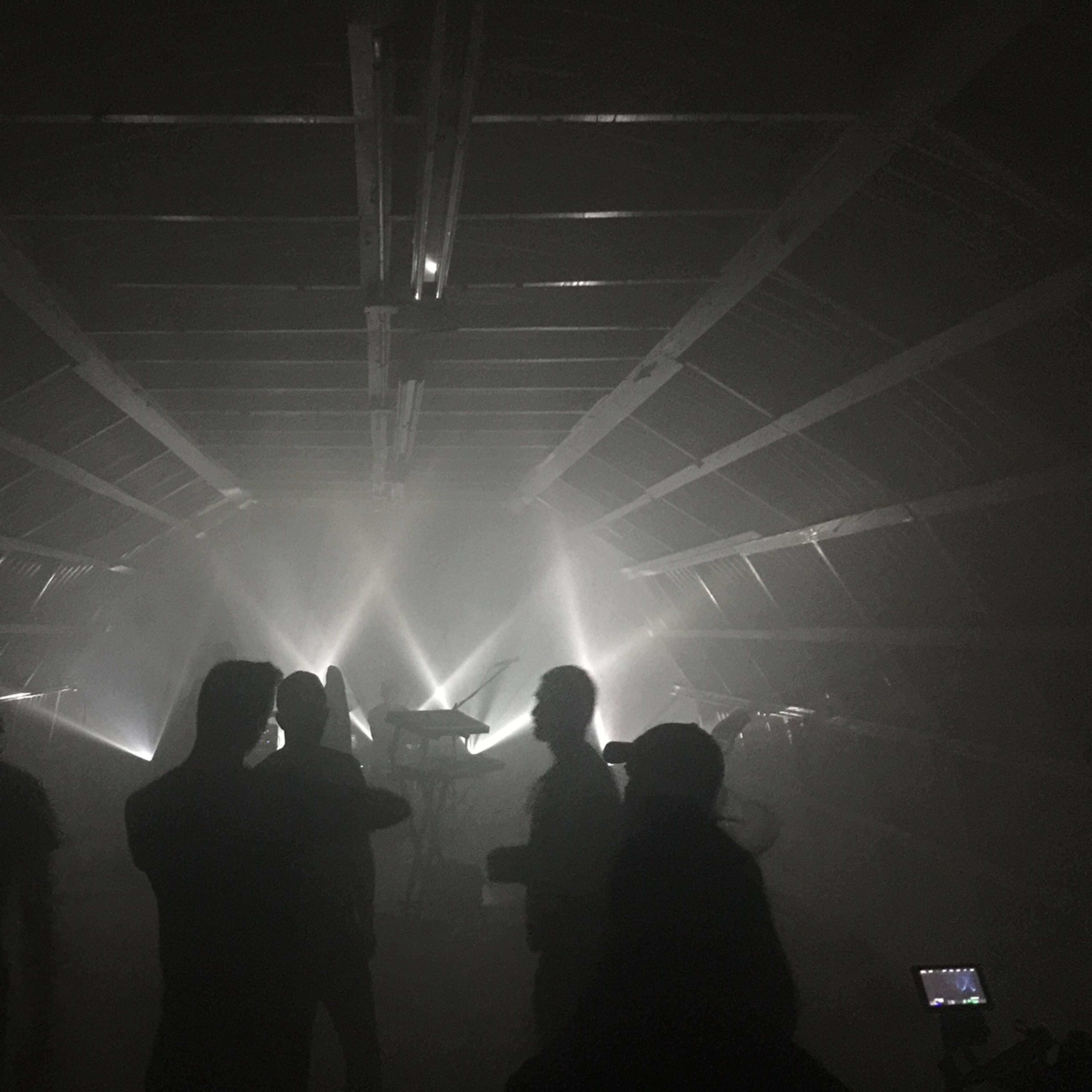 A group of people stands in a dimly lit space, with beams of light cutting through a hazy atmosphere and musical instruments visible in the background.