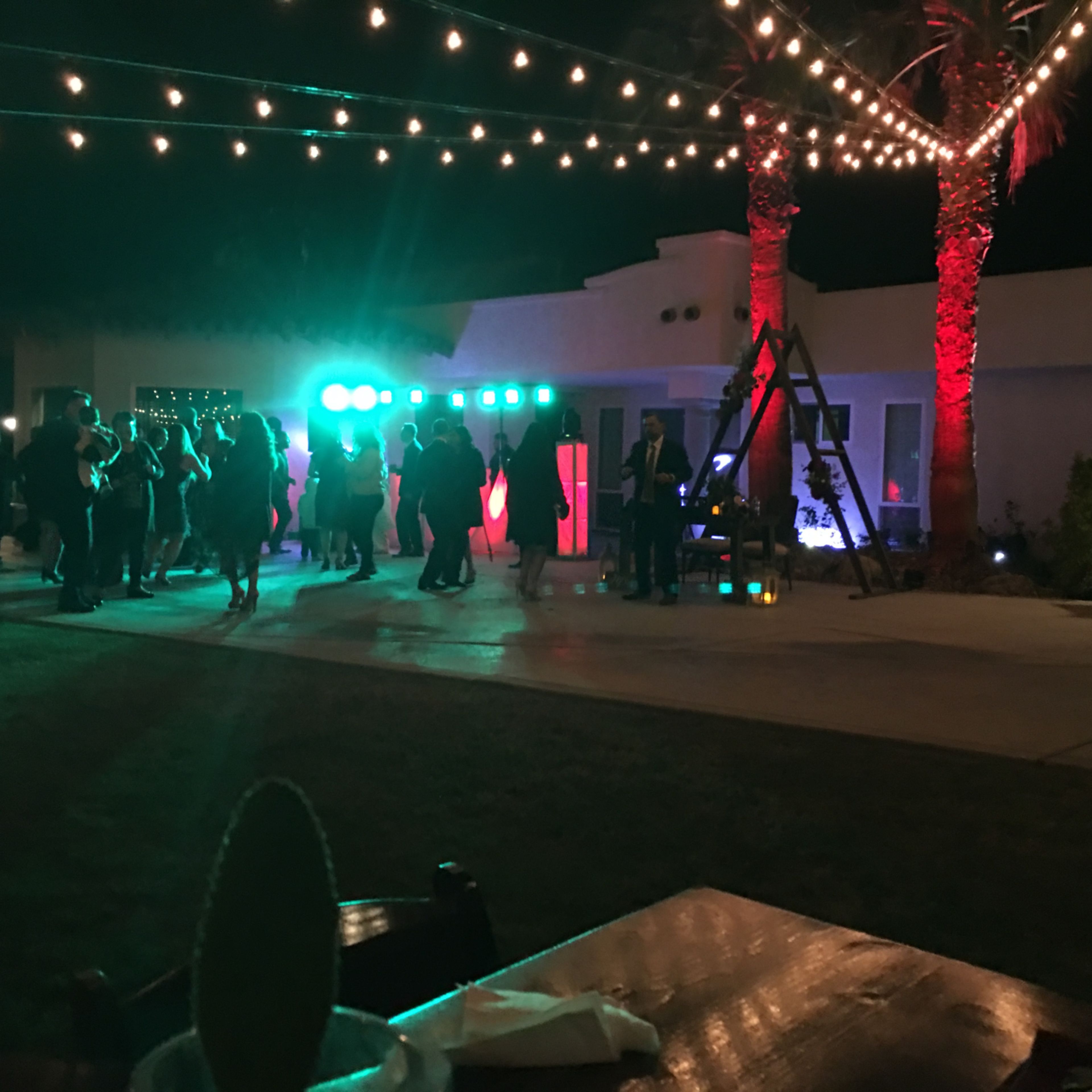 A lively outdoor event features guests dancing under string lights, with colored lighting illuminating the scene.