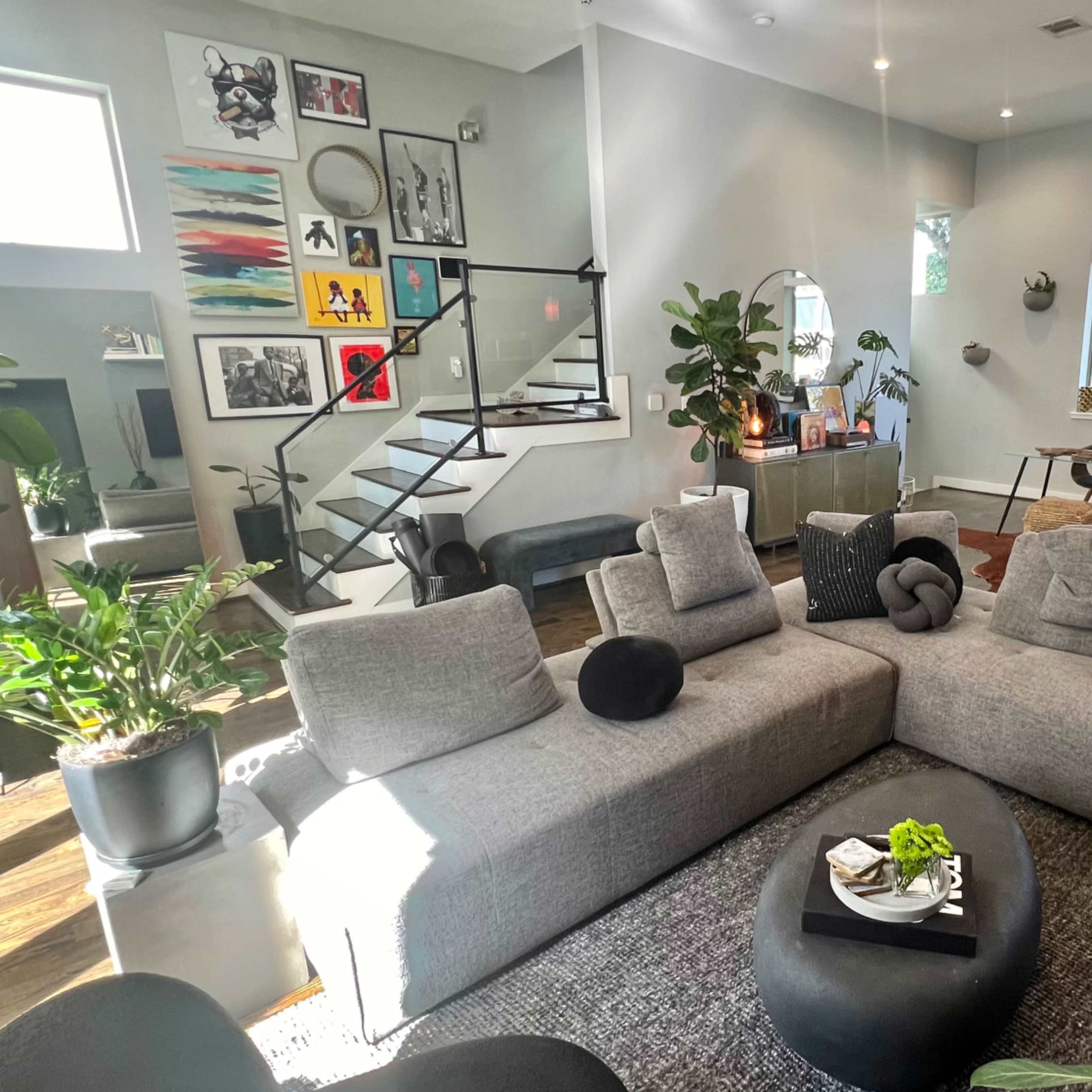 A modern living room with a gray sectional sofa, various plants, a staircase, and artwork displayed on the walls.