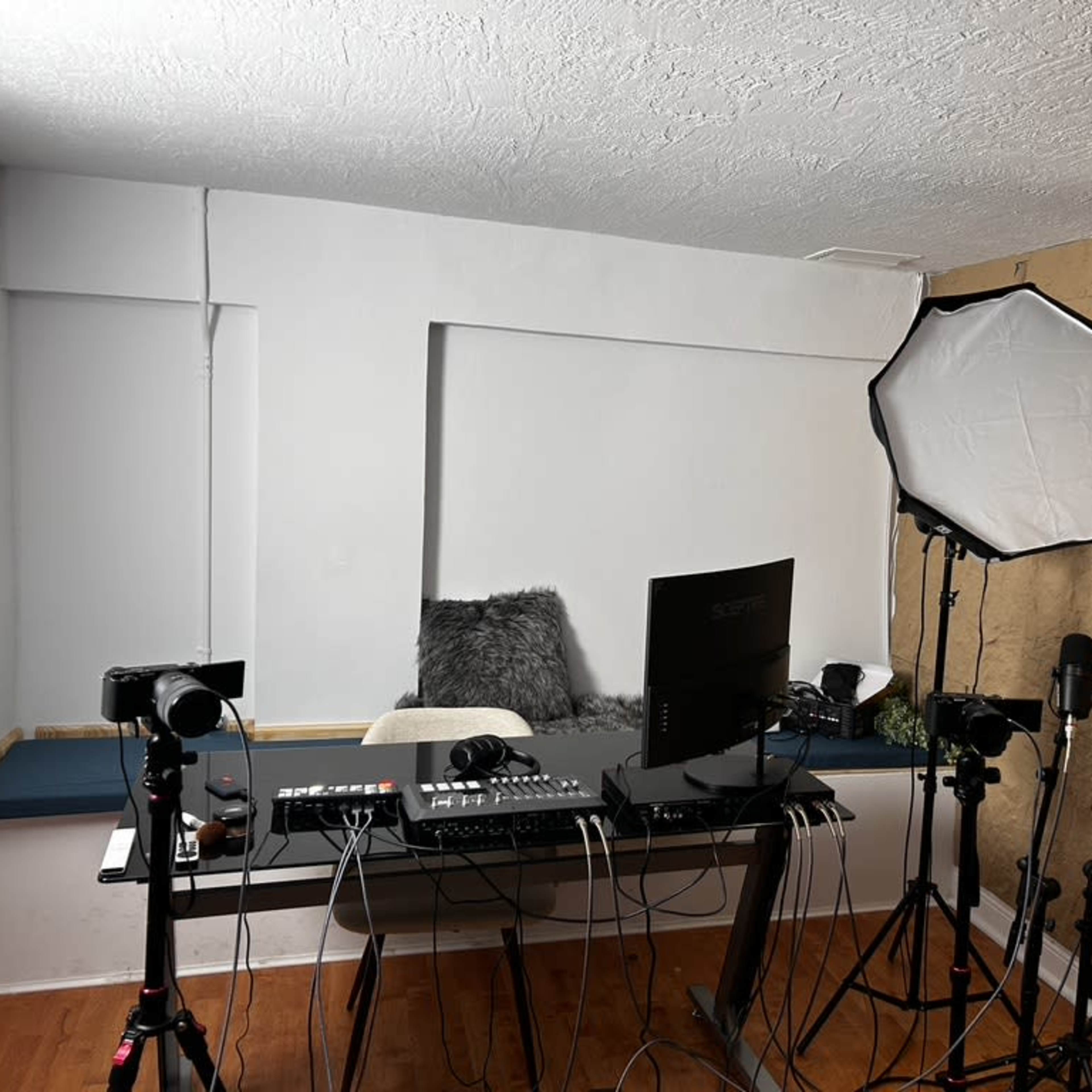 A recording studio setup featuring a large desk with a computer, multiple cameras, microphones on stands, and a softbox light.