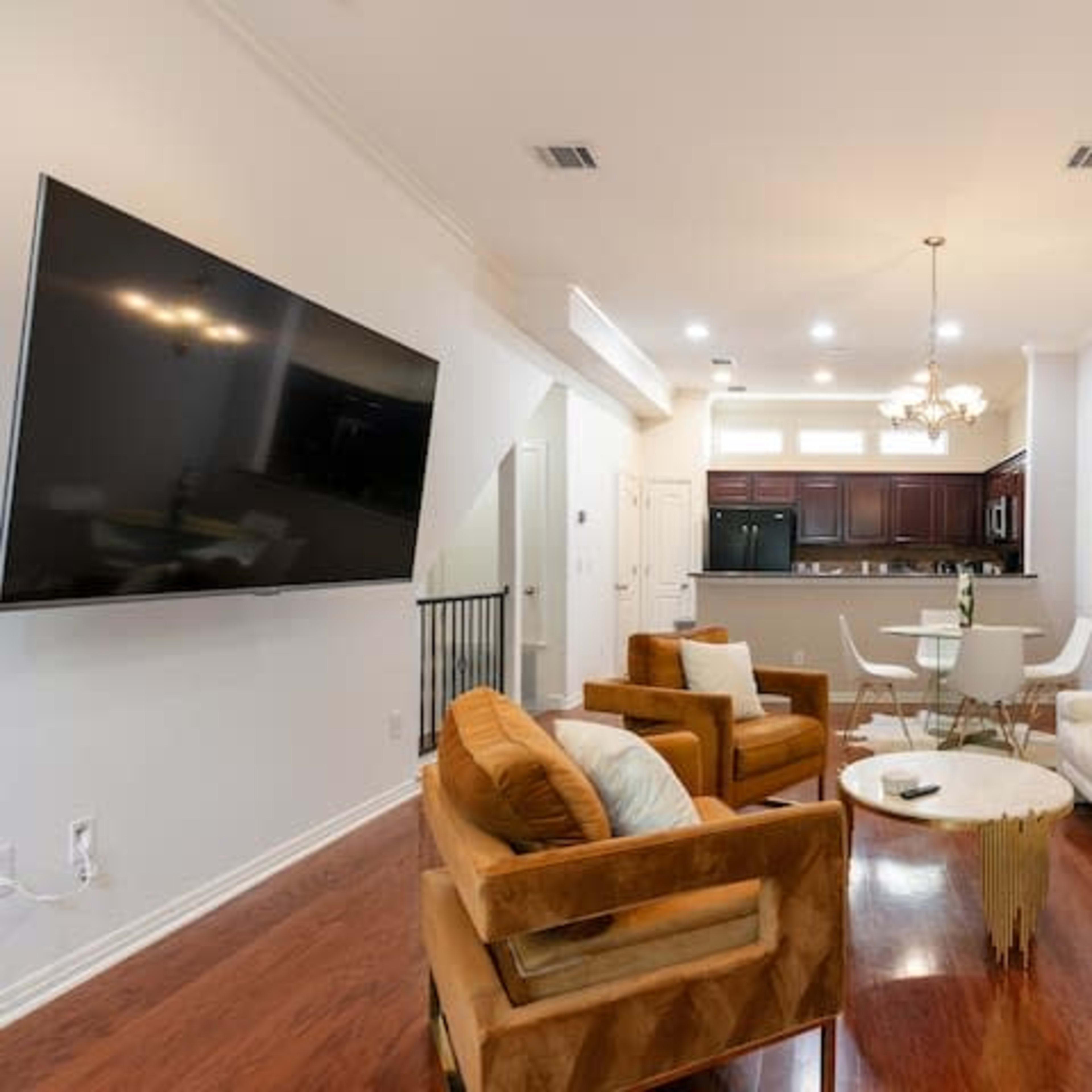 A living room with a large television mounted on the wall, a white sectional sofa, two brown armchairs, a circular coffee table, and a view into a kitchen area with dark wood cabinets.
